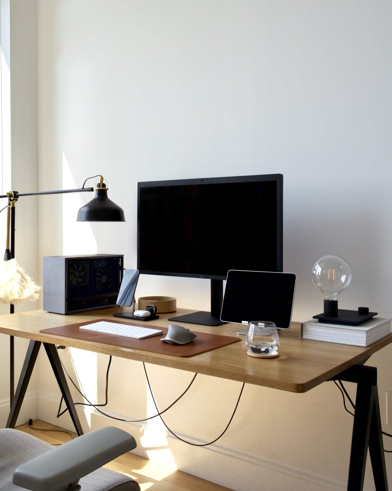A minimalistic desk setup with a modern LG monitor, tablet stand, keyboard on a leather mat, designer lamp, and decorative items, including books and a glass orb, all bathed in natural light