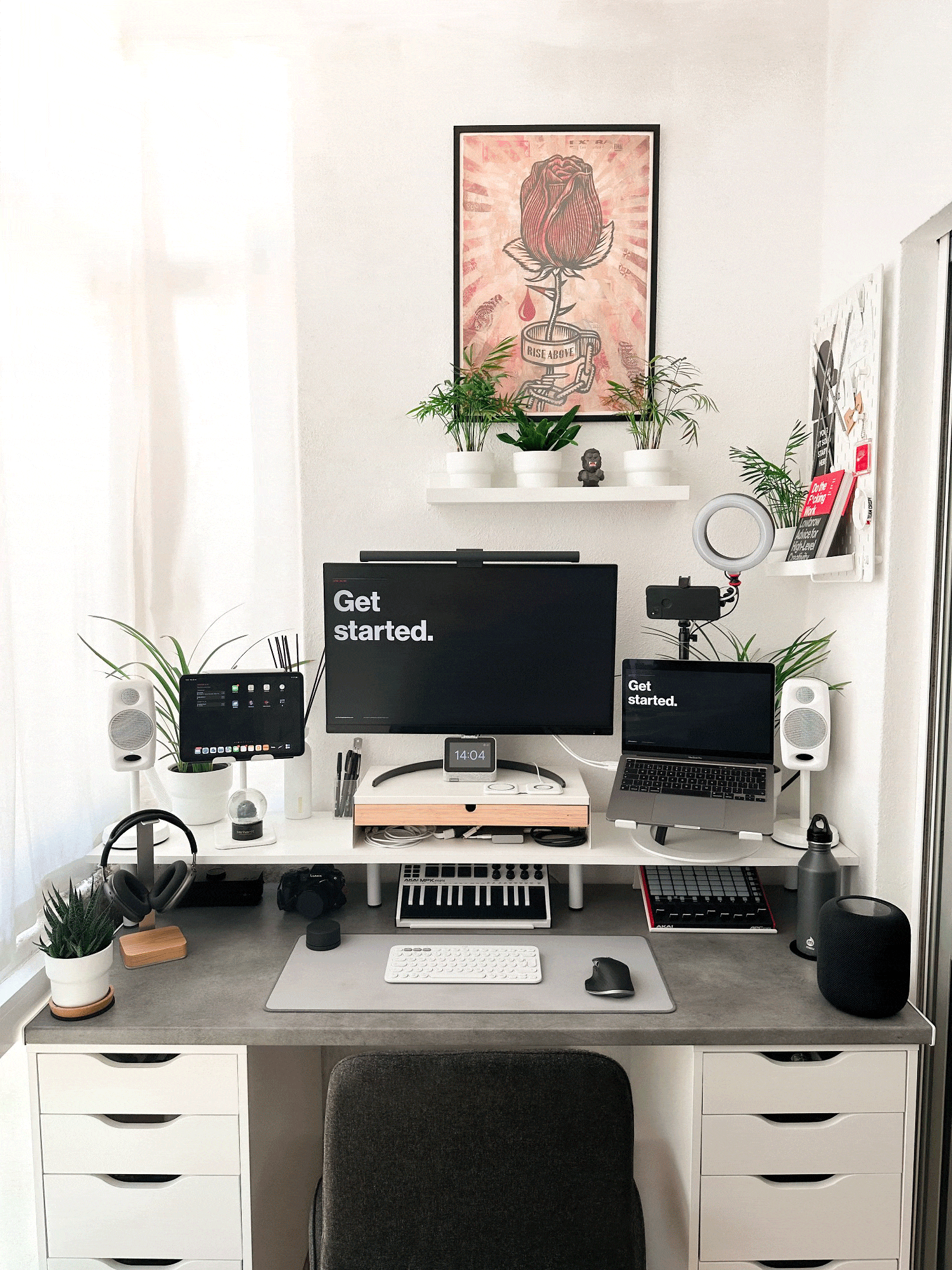 A minimalist desk setup with a laptop on a stand, a MIDI keyboard, a camera, headphones, a microphone, and assorted plants, with a motivational poster on the wall, all in a clean, bright space