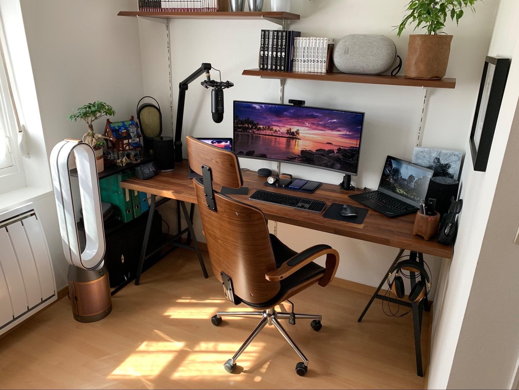 A well-organised desk setup with a wooden desk and chair, dual monitors, laptop, microphone, headphones, and various desk accessories in a room with natural lighting
