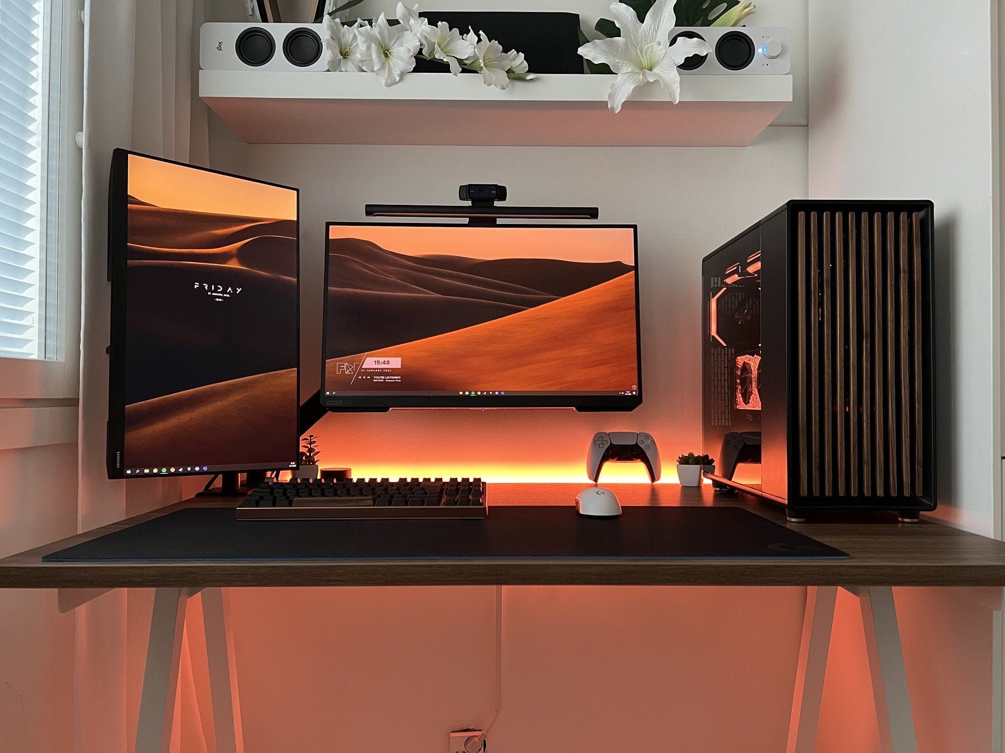 A sleek desk setup featuring two monitors with matching wallpapers, a high-end PC tower, mechanical keyboard, and mouse, with decorative plants and speakers under soft ambient lighting