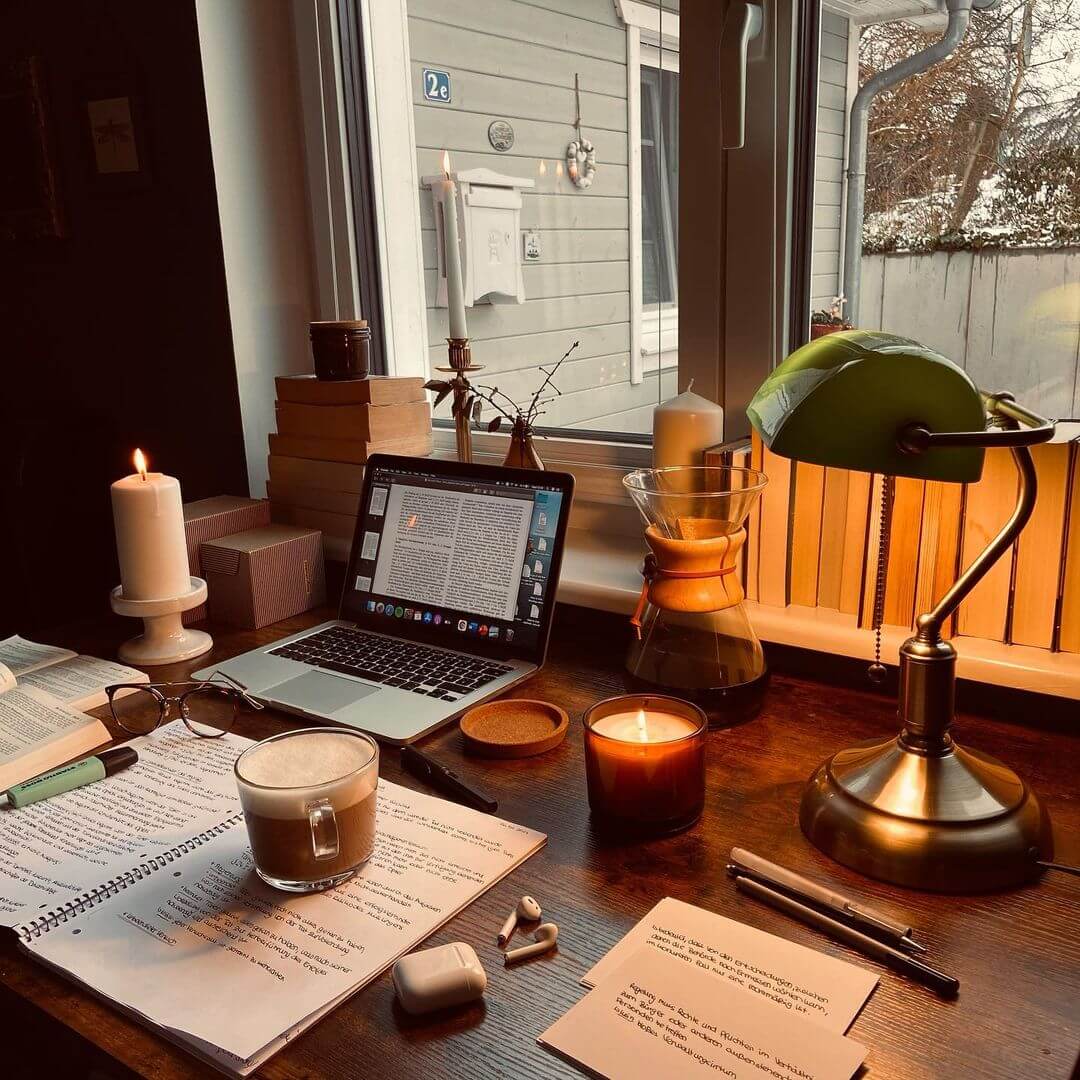 A cosy study setup with an open laptop, handwritten notes, glasses, lit candles, a cup of coffee, and a green desk lamp by a window