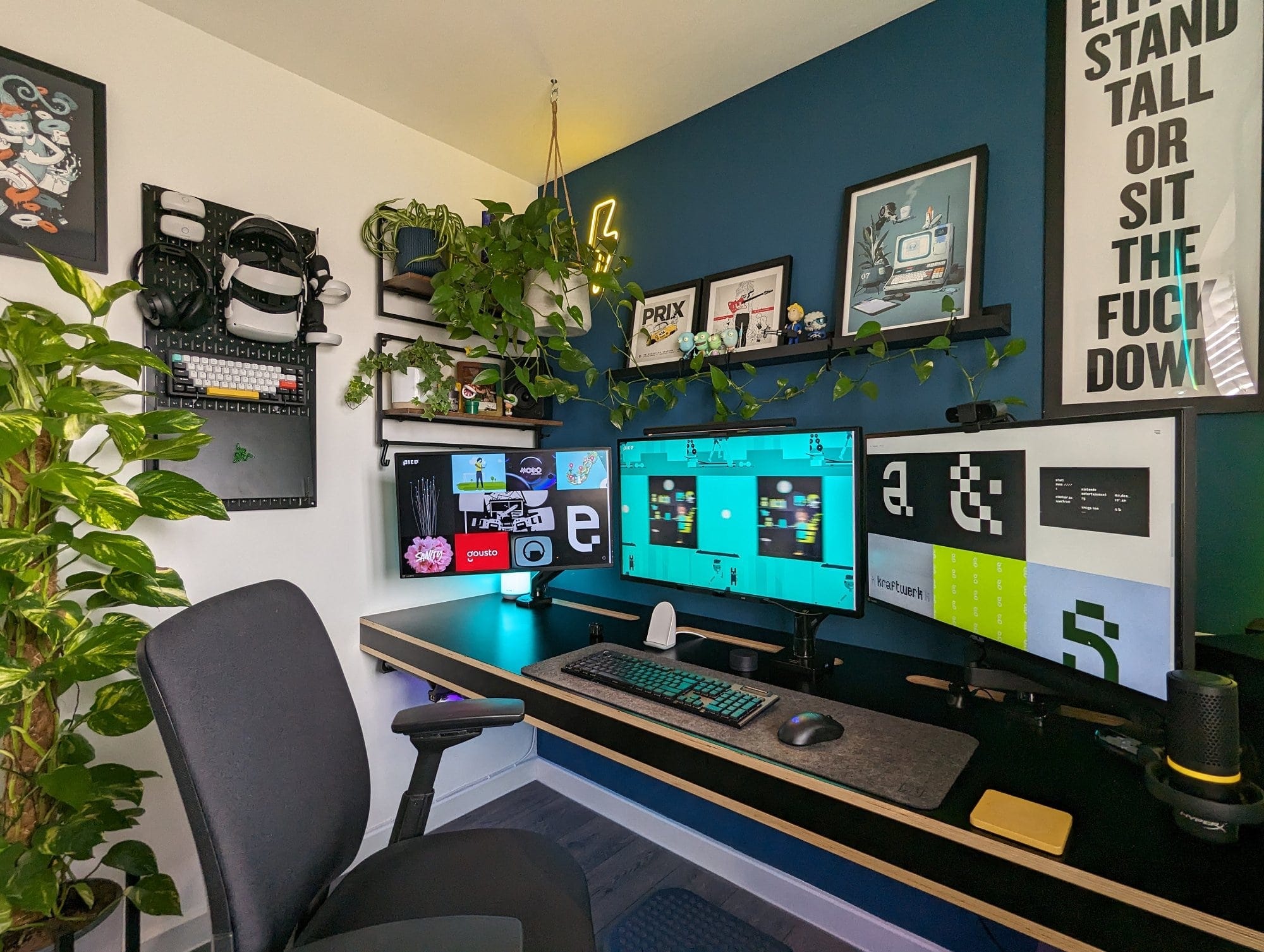 A modern home office setup with a triple-monitor desk, accented by lush green plants, a pegboard organiser with tech accessories, framed art, and motivational posters, illuminated by stylish lighting