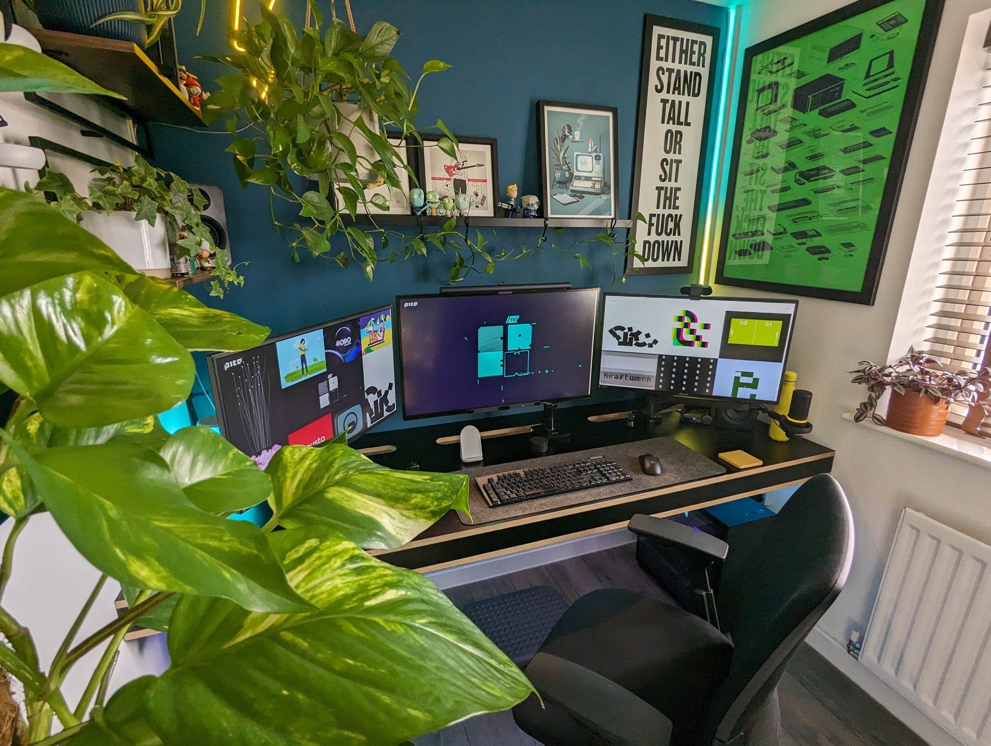 A modern home office setup featuring a triple-monitor desk, lush green plants, framed artwork, motivational posters, and ambient LED lighting