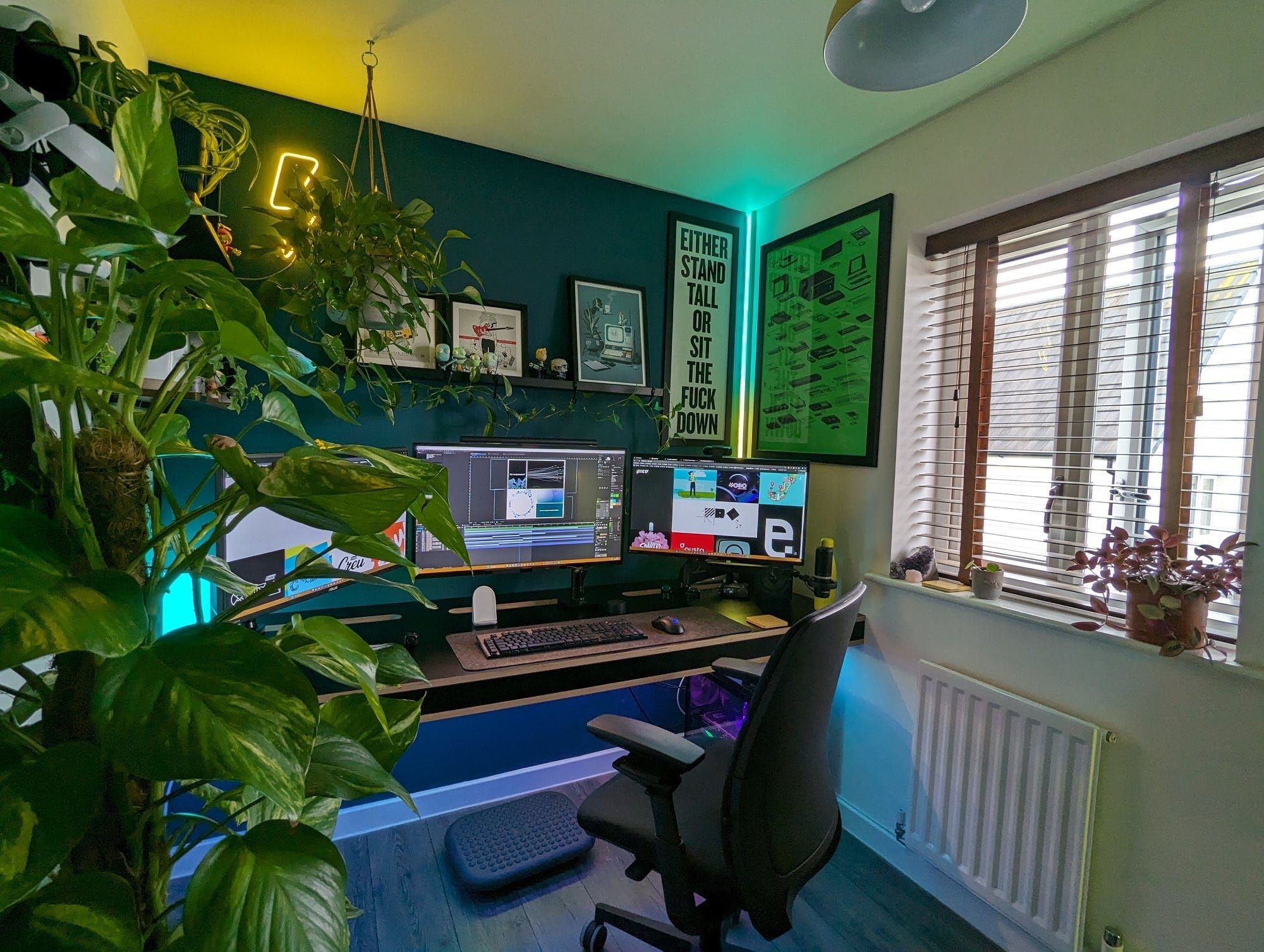 A contemporary home office featuring a triple-monitor desk setup, surrounded by lush plants, motivational posters, and modern lighting, with natural light streaming in from a nearby window