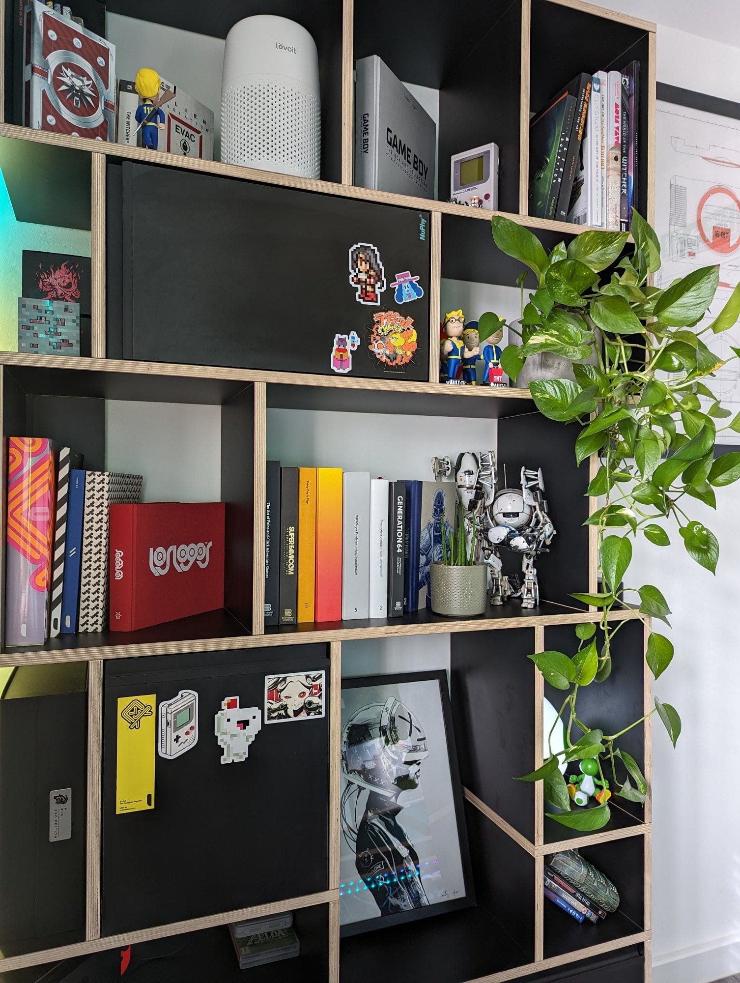 A bookshelf filled with books, tech gadgets, collectibles, and potted plants, featuring a mix of retro and modern items, decorated with stickers and framed artwork, and accentuated by a hanging plant
