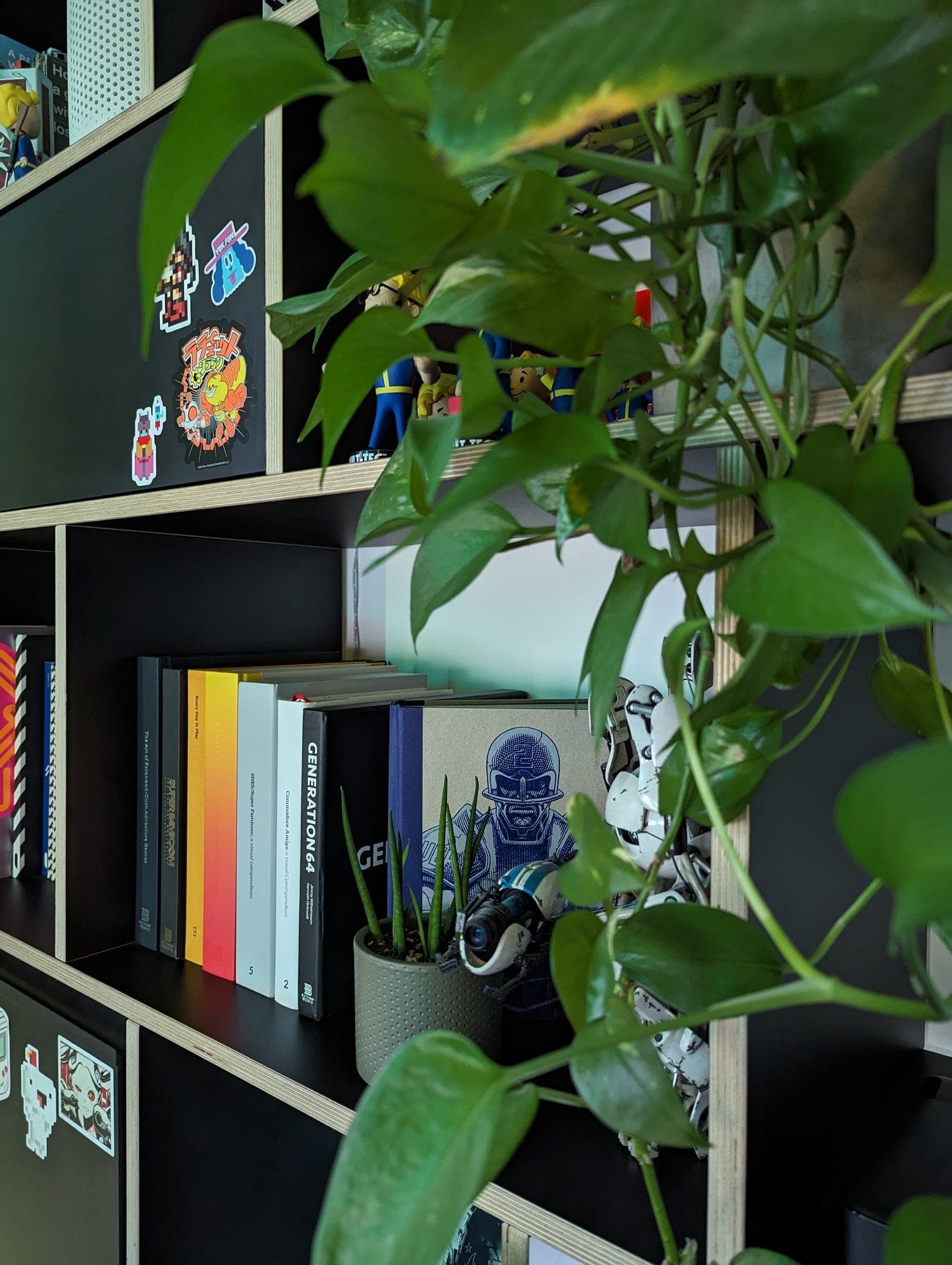 A bookshelf adorned with books, action figures, and decorative stickers, partially obscured by vibrant green foliage from a nearby plant