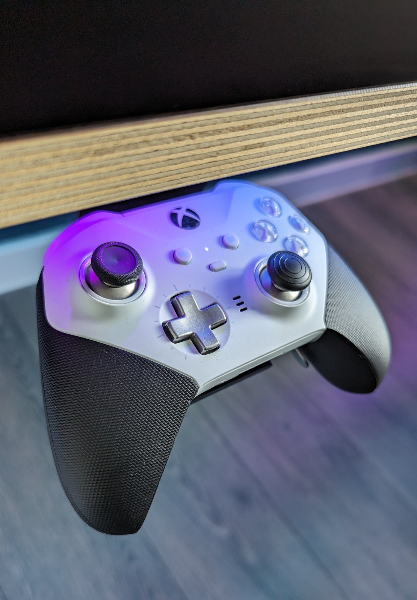A close-up of an Xbox Elite Series 2 wireless controller stored under a desk, illuminated by ambient purple lighting