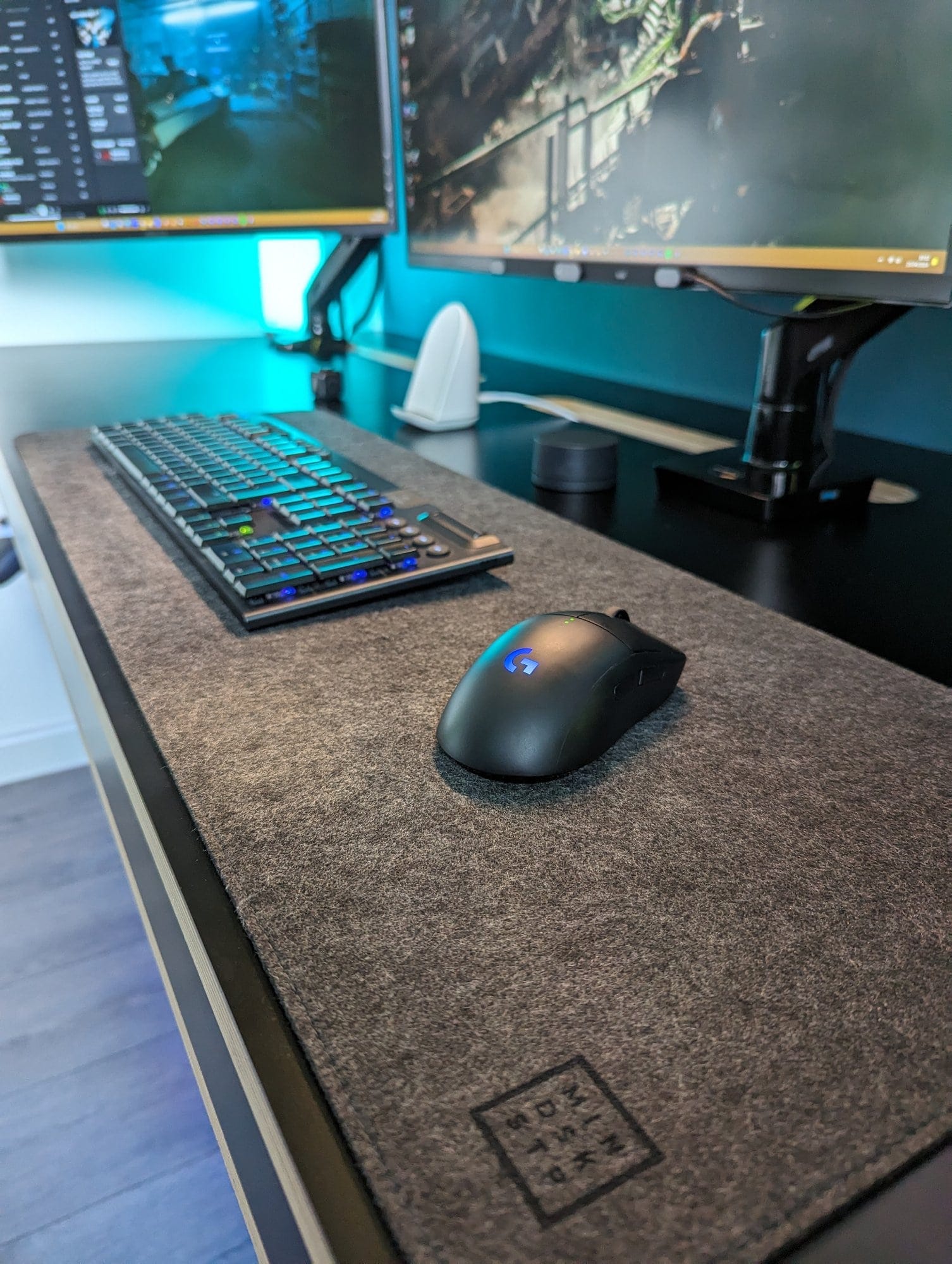 A close-up of a modern home office desk featuring a mechanical keyboard and a wireless mouse on a grey felt desk mat, with multiple monitors displaying game graphics in the background