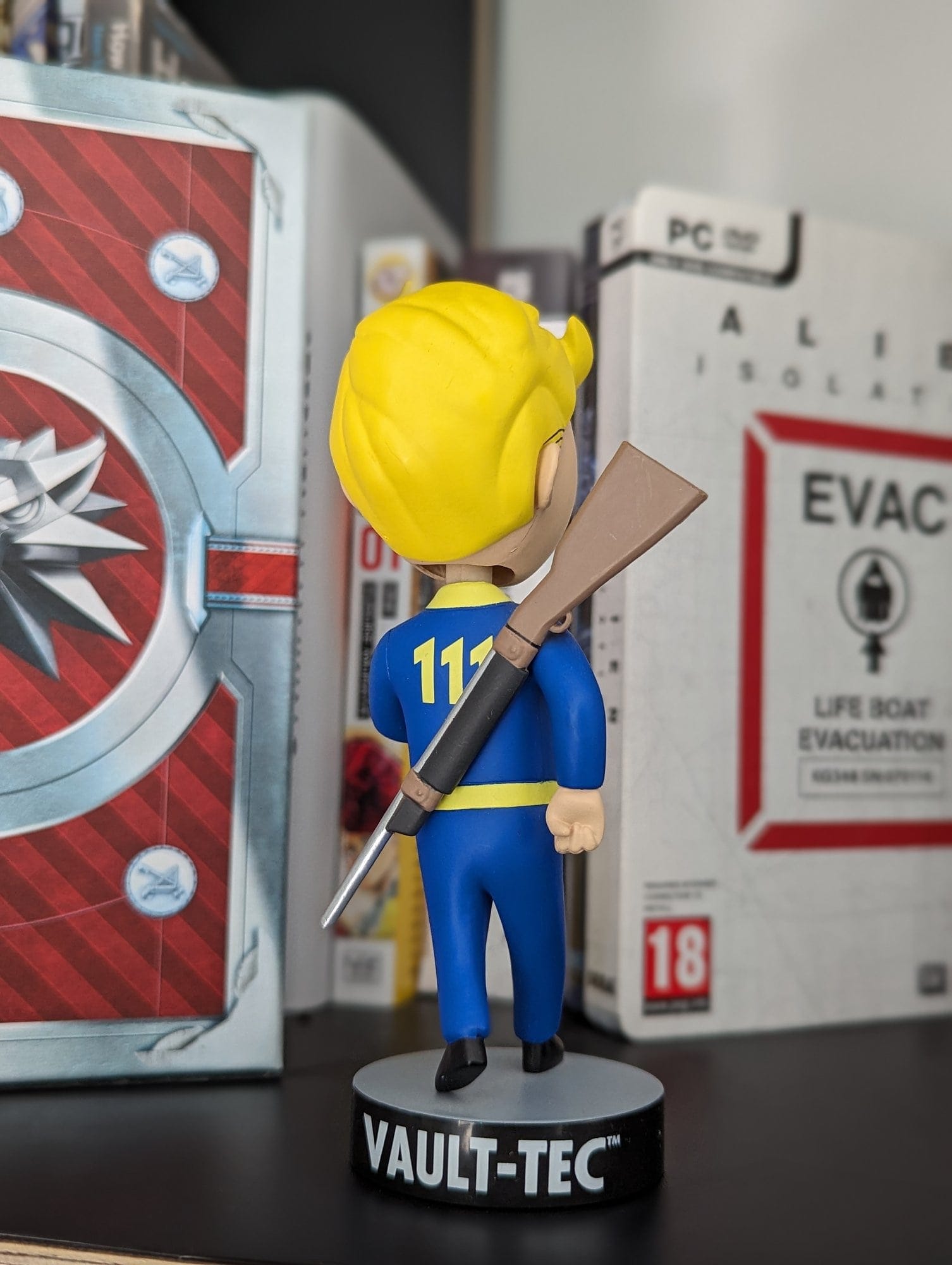 A Vault-Tec bobblehead figurine from the Fallout video game series is displayed on a shelf, with video game boxes in the background