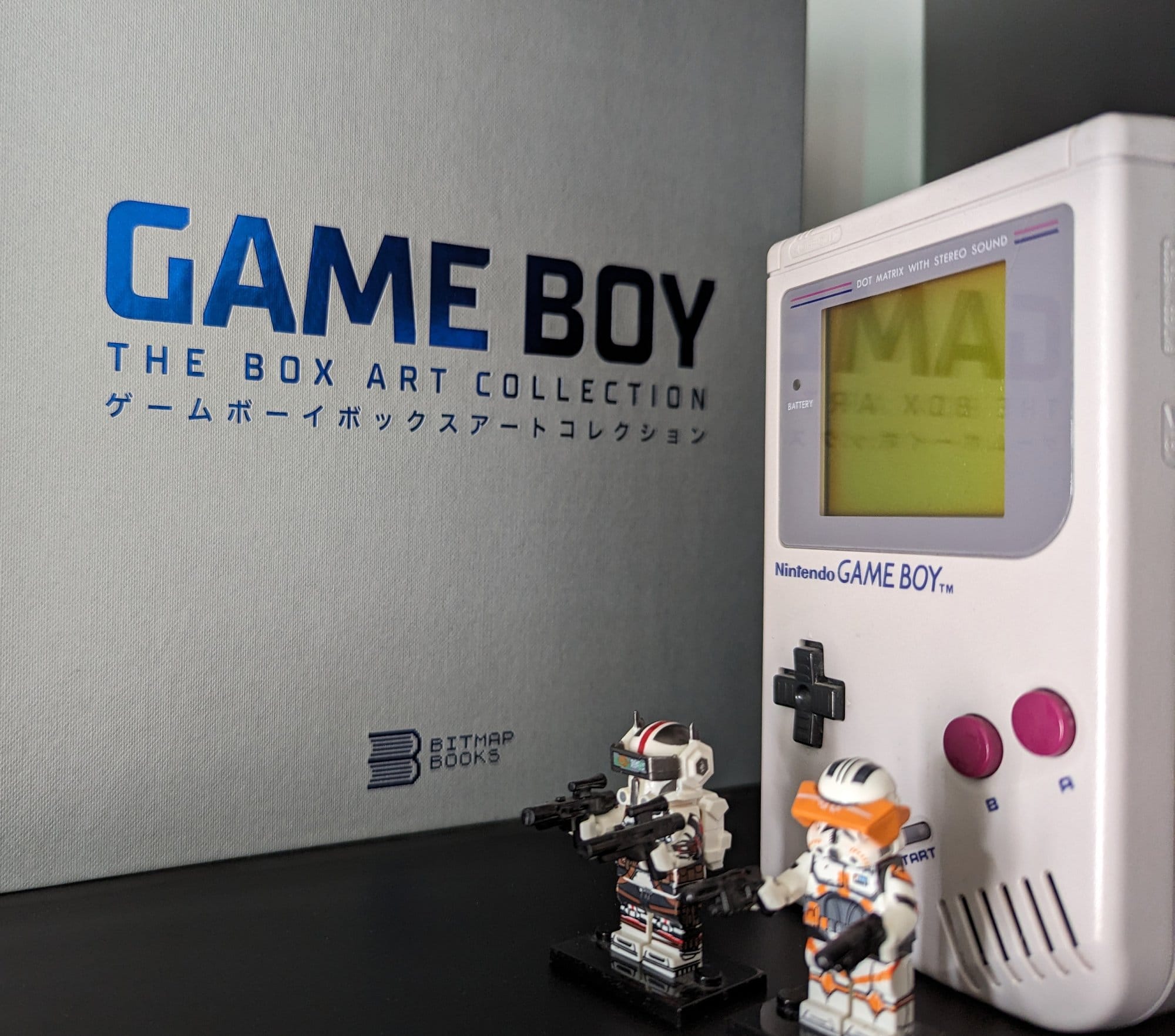 A close-up of a vintage Nintendo Game Boy displayed alongside a “Game Boy: The Box Art Collection” book, with two small action figures positioned in front