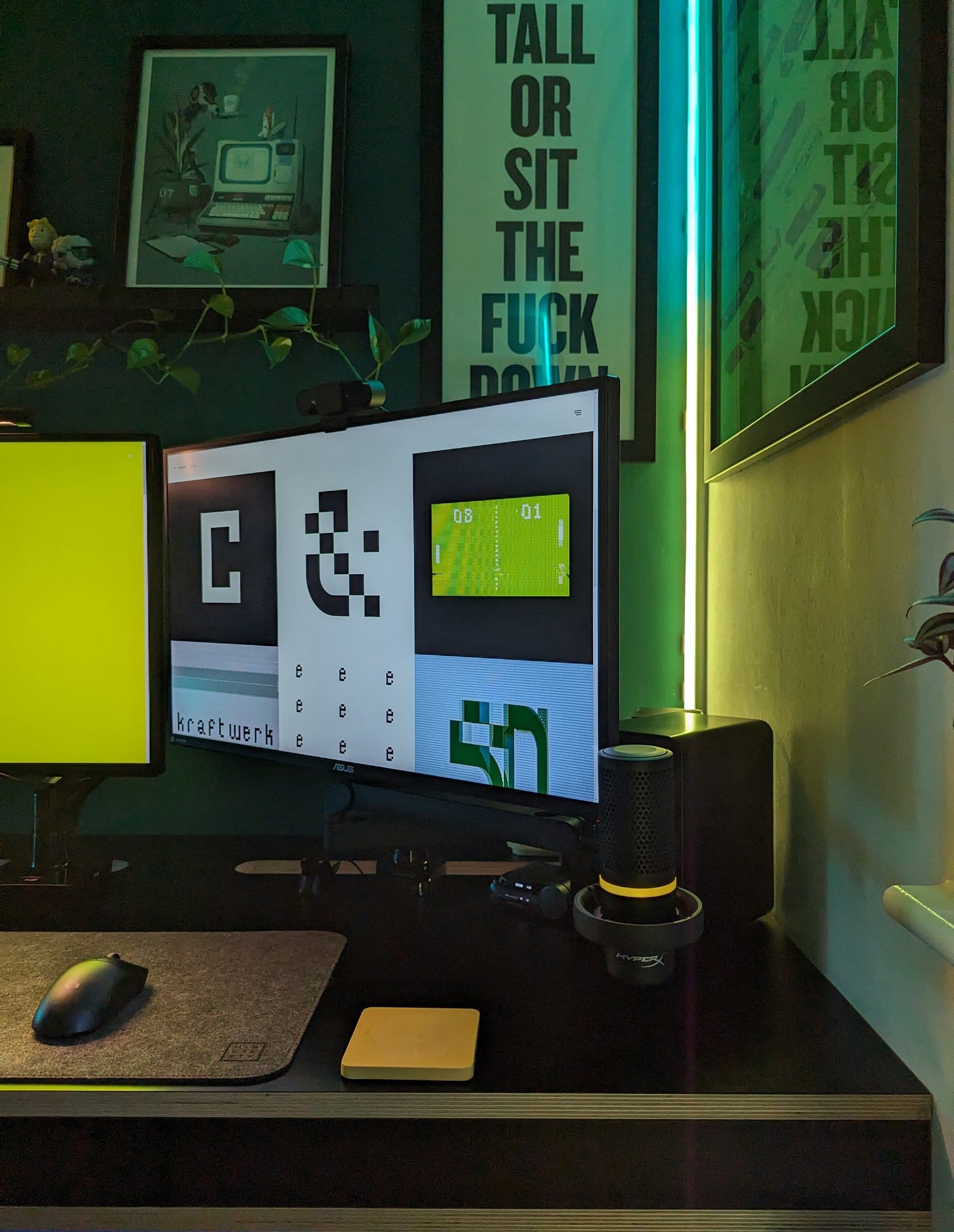 A close-up of a home office desk setup featuring a triple-monitor display, with colourful lighting, framed art, and motivational posters in the background, alongside a mouse and a microphone on the desk