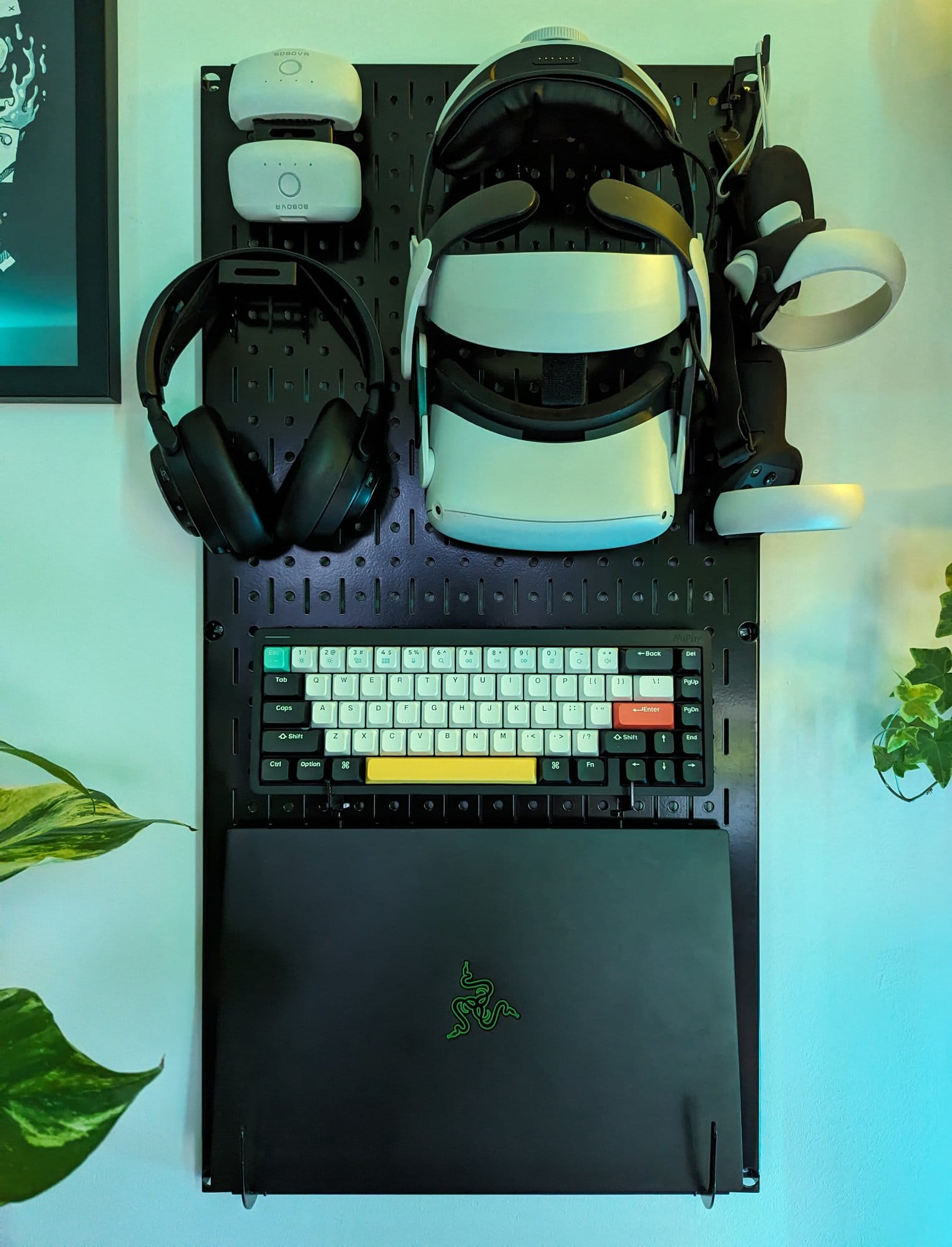 A pegboard organiser mounted on a wall, holding a VR headset, controllers, headphones, a mechanical keyboard, and a Razer laptop, with nearby green plants adding a touch of nature