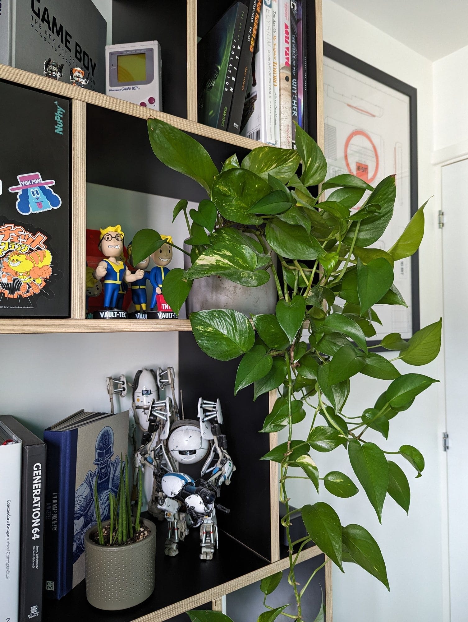 A Tylko bookshelf filled with books, collectibles, and a potted plant, featuring a retro Game Boy, Vault-Tec figures, and a robot model from Portal, with leafy green vines adding a touch of nature