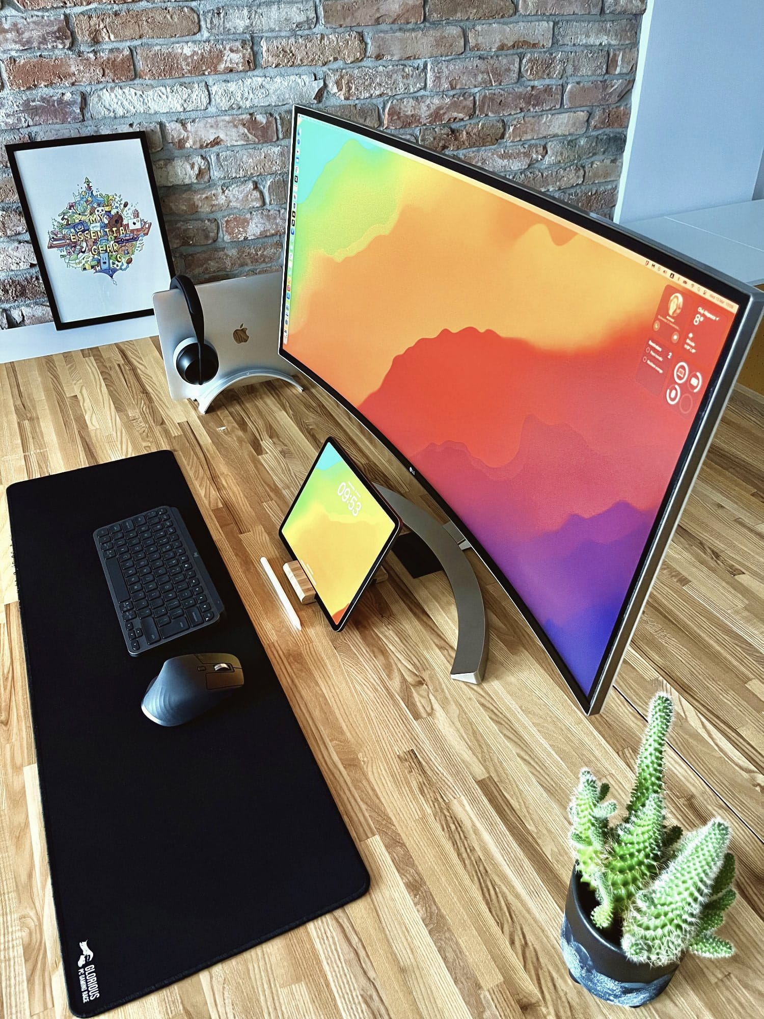 A contemporary office setup with a large curved monitor displaying vibrant wallpapers, an Apple desktop computer, wireless keyboard and mouse on a black desk pad, a framed colourful artwork, and a potted cactus on a wooden desk against a brick wall