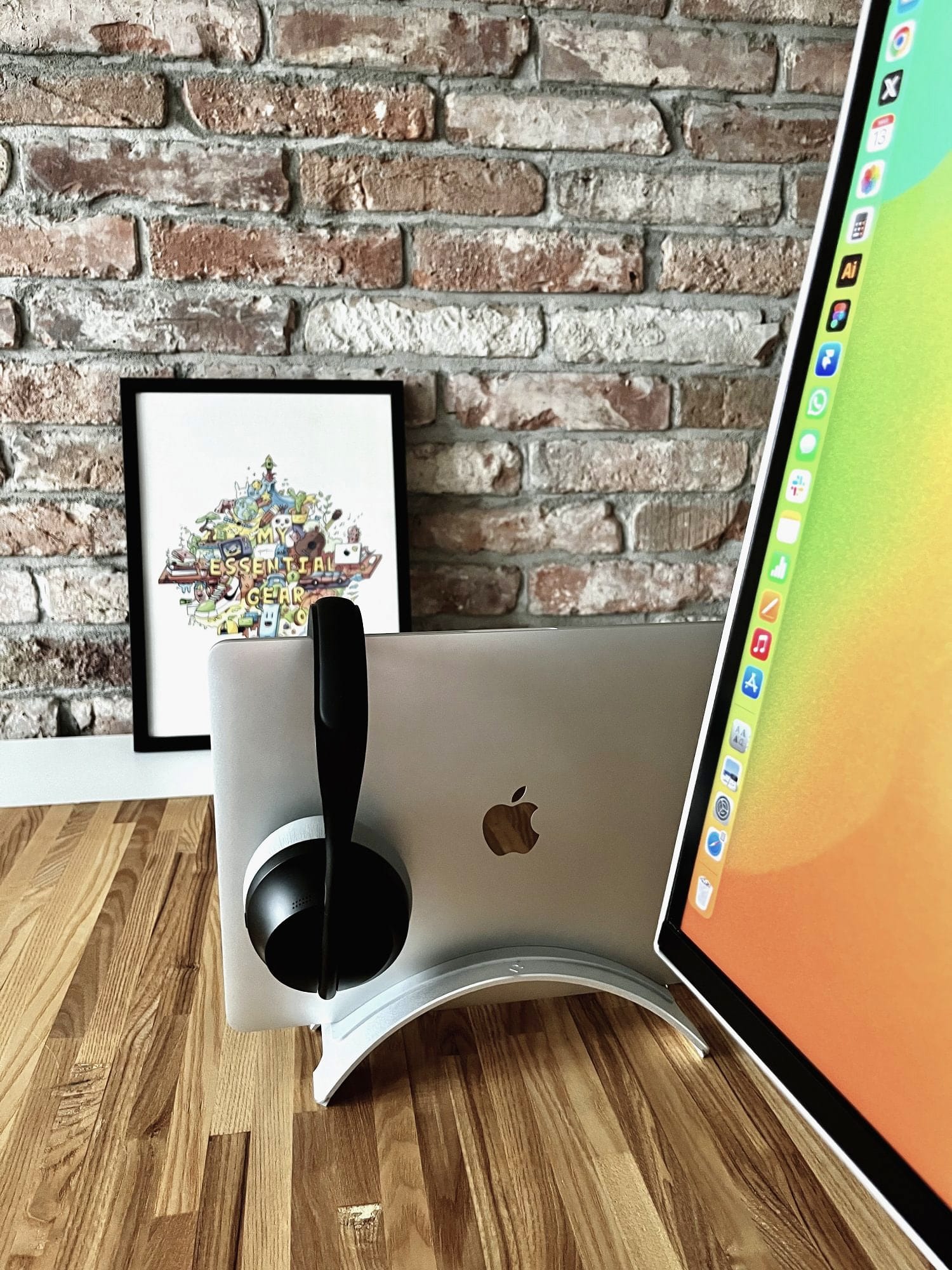 A corner of the modern desk setup with a monitor, a MacBook on a stand, headphones, framed poster, and exposed brick wall