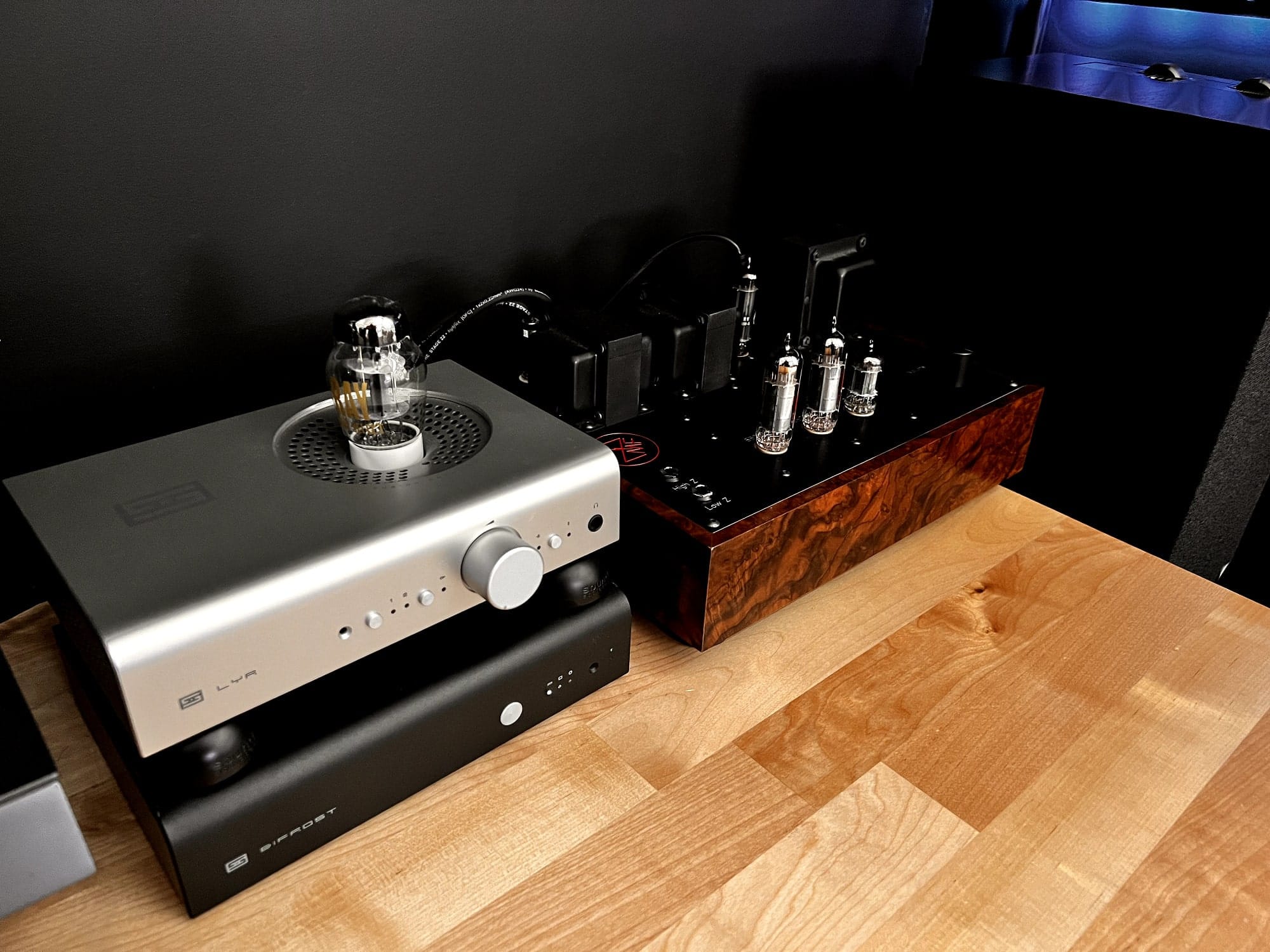 A close-up of high-end audio equipment on a wooden desk, featuring a silver Schiit Lyr 3 tube amplifier stacked on a black Schiit Bifrost DAC, and a custom Feliks Audio Euforia tube amplifier with a wooden enclosure and exposed vacuum tubes.