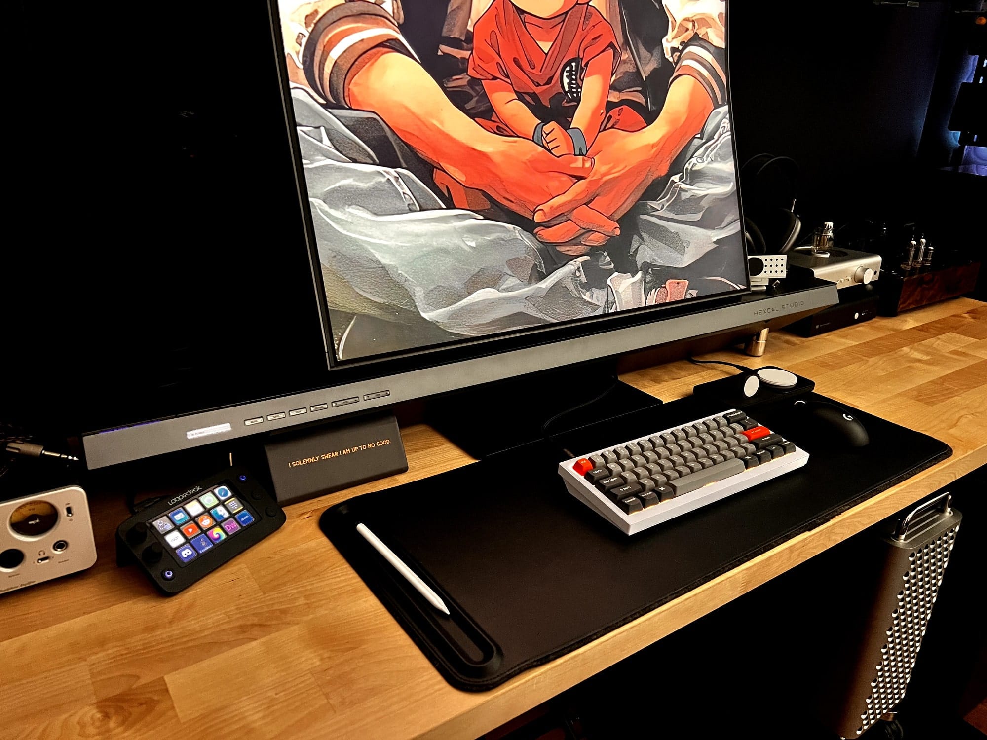 A modern desk setup featuring a large curved monitor displaying anime, a grey Keychron Q4 mechanical keyboard with custom keycaps, a Logitech G Pro wireless gaming mouse, a Stream Deck, and an Apple Pencil on a black desk mat, with high-end audio equipment and a Mac Pro tower placed underneath the desk