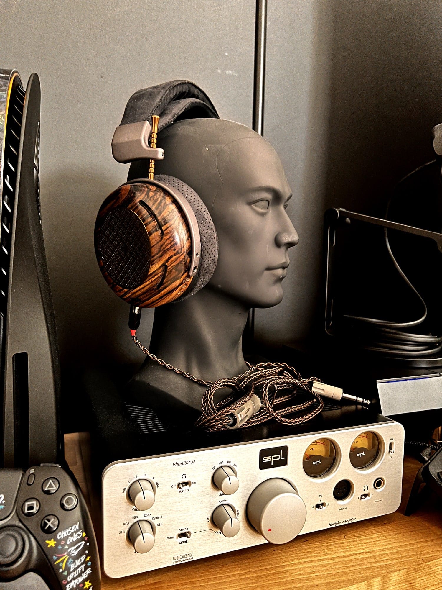 A close-up of a wooden-cup headphone on a mannequin head next to the Phonitor XE headphone amplifier and a gaming console controller in the foreground