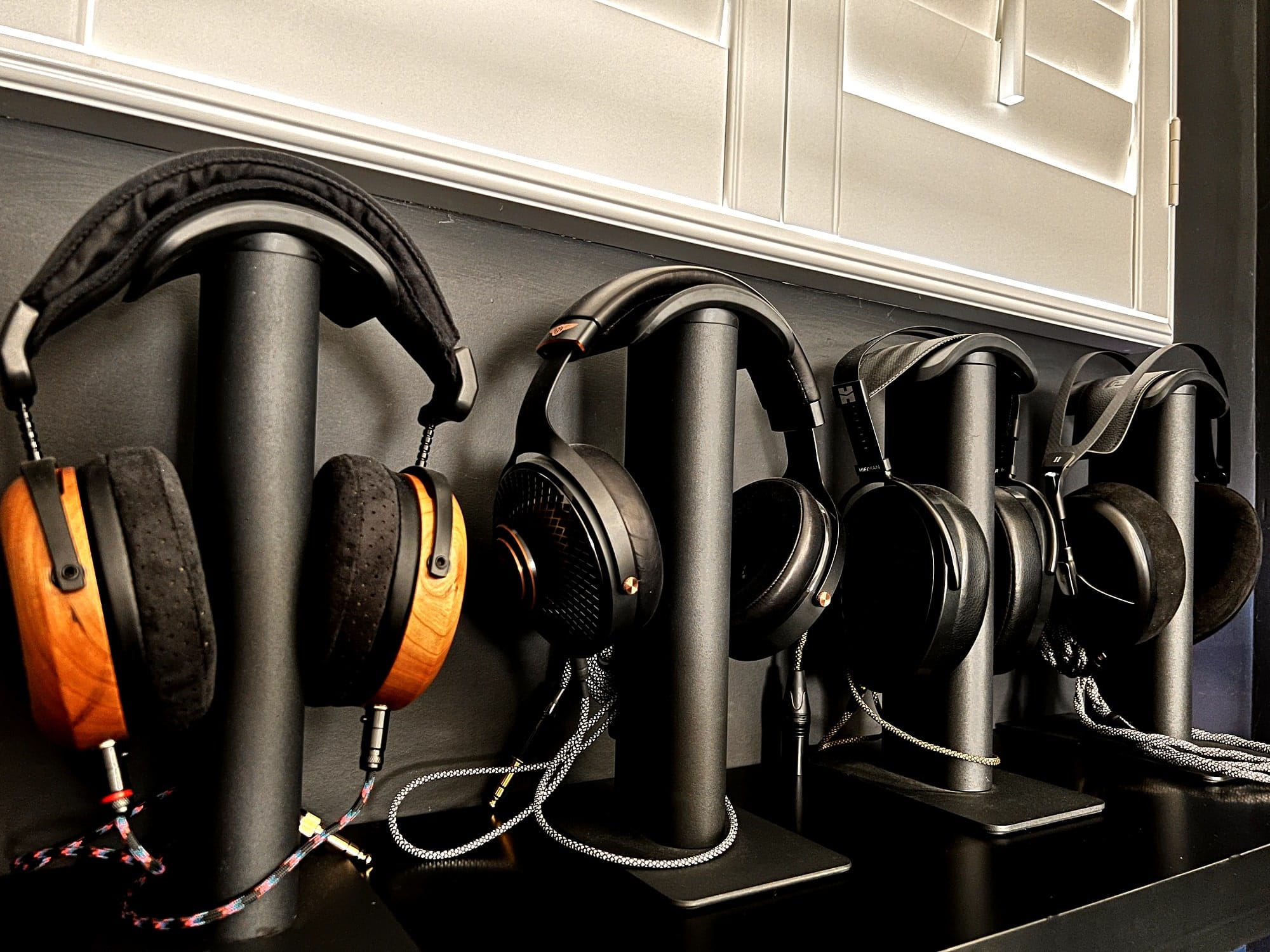 A collection of high-end headphones displayed on stands, featuring the ZMF Caldera, ZMF Atrium, Meze Empyrean Elite, and HiFiMan Arya Stealth, all arranged neatly under a window with white shutters
