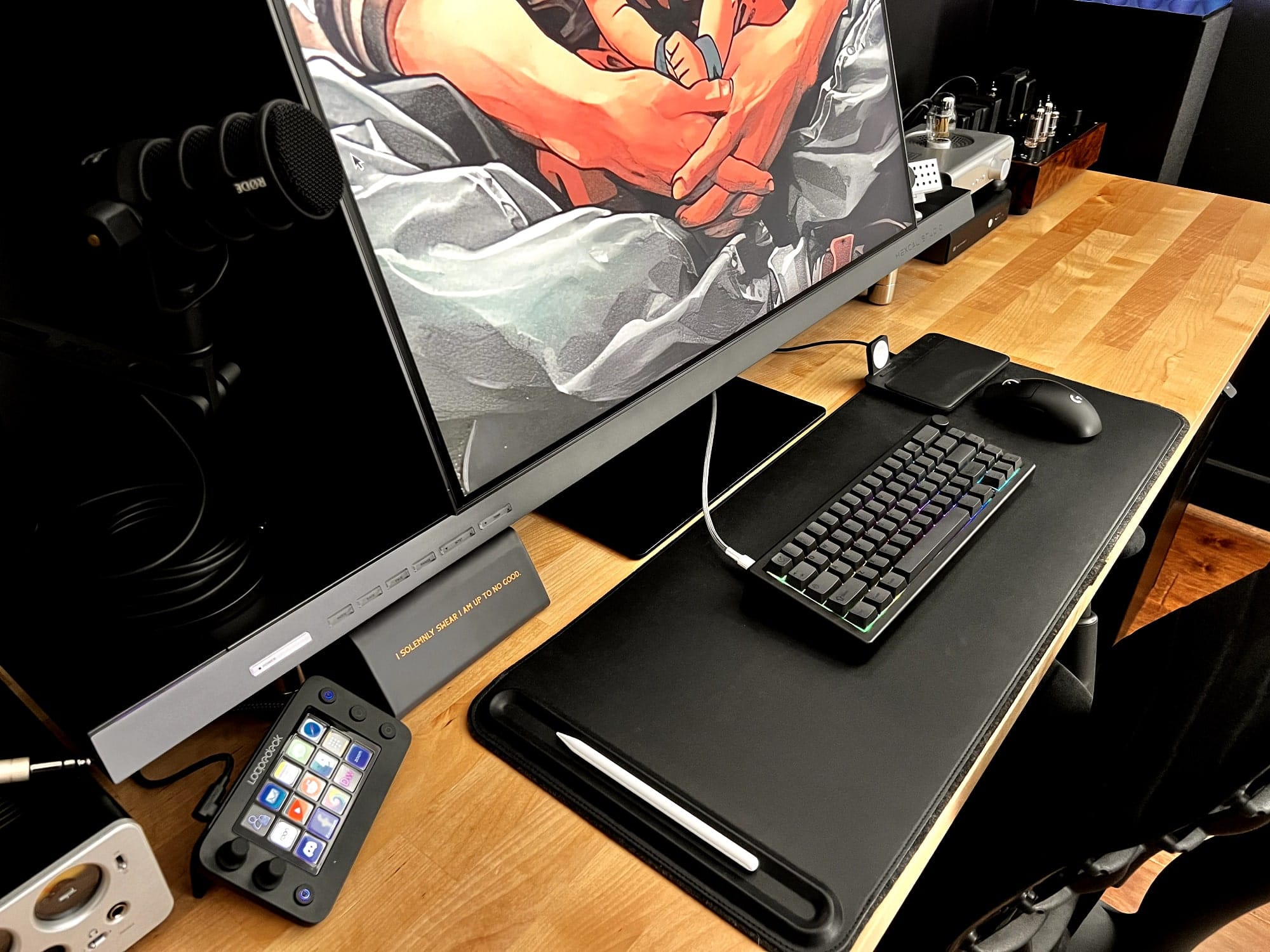 A close-up view of a tidy gaming desk setup, featuring a large Samsung Odyssey Ark 2 monitor with an anime display, a mechanical keyboard, a gaming mouse, a Stream Deck, and a RØDE microphone, all arranged neatly on a wooden surface