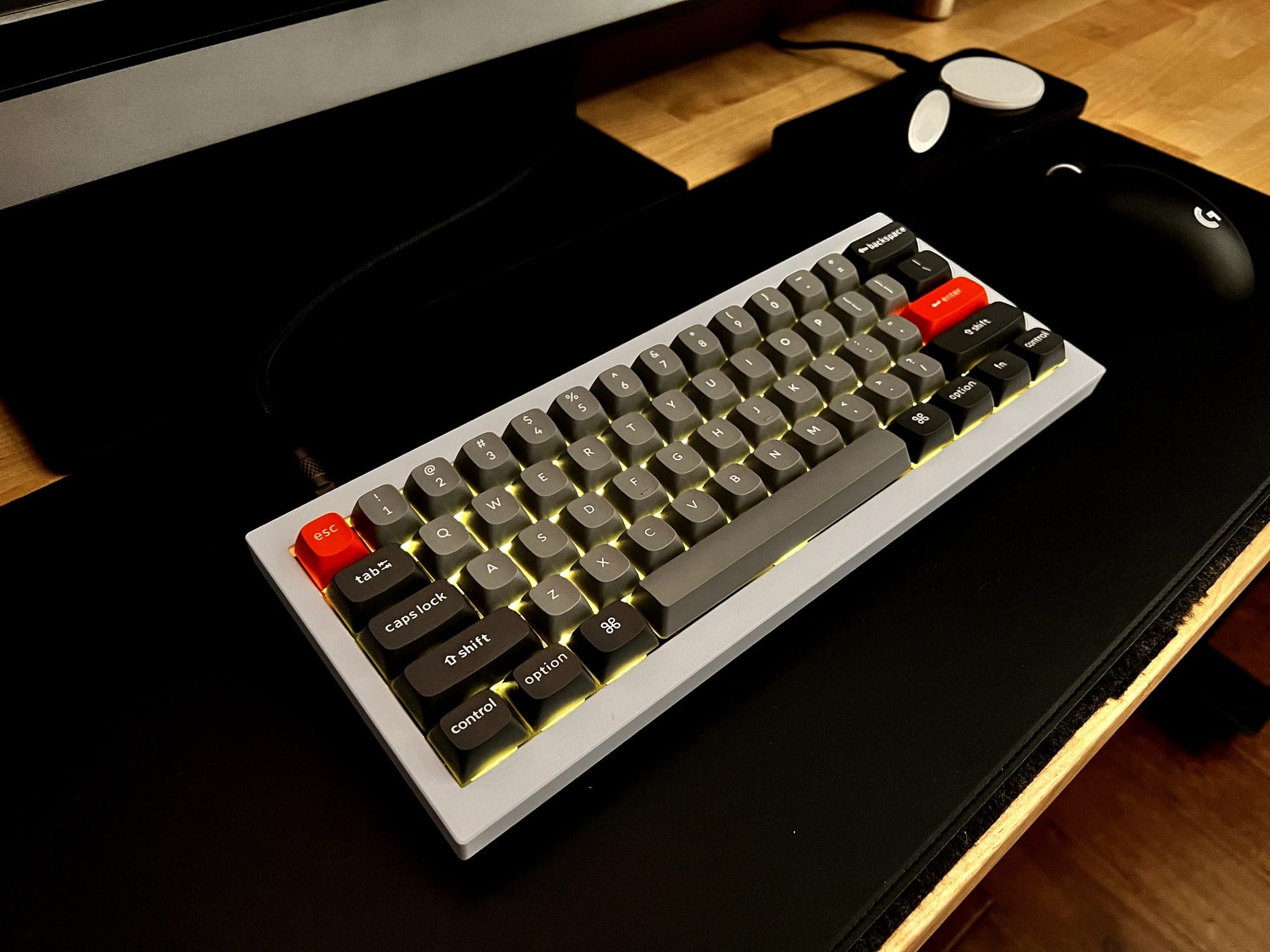 A close-up of a grey Keychron mechanical keyboard with custom keycaps, including an orange escape key and red return and shift keys, backlit with yellow LEDs, placed on a black desk mat next to a Logitech G Pro wireless gaming mouse