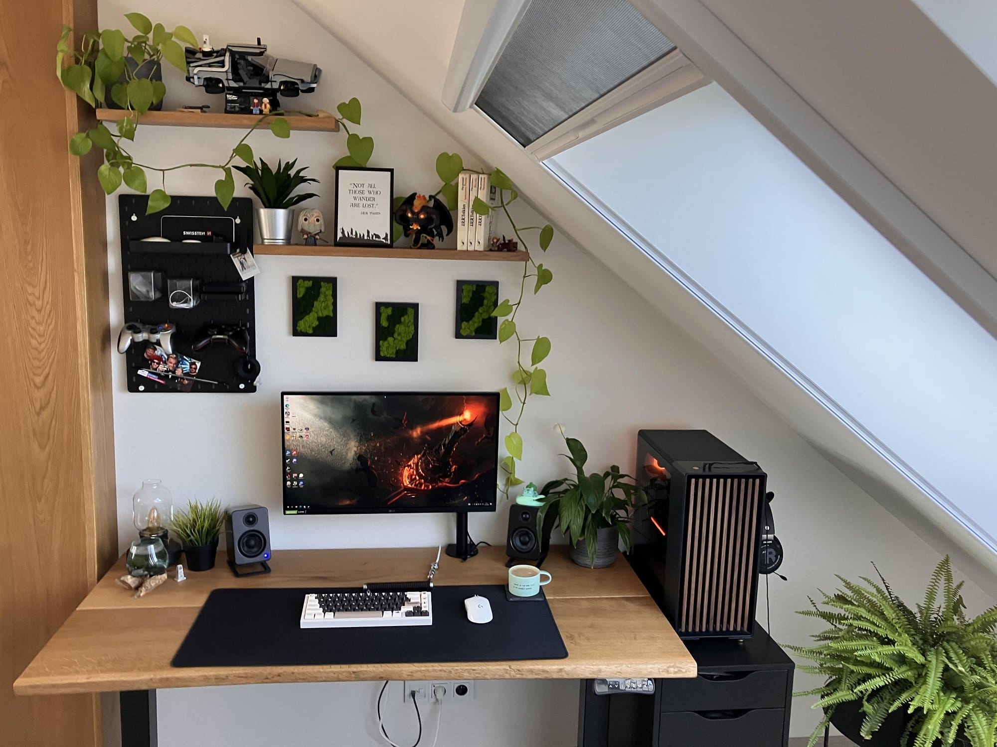 A neatly organised home office setup with a wooden desk, computer, and multiple plants, illuminated by natural light from a slanted window, featuring shelves with decor and greenery