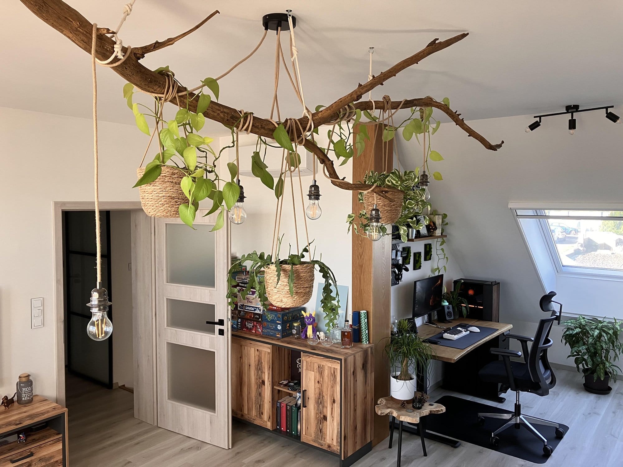 A modern home office featuring a wooden desk with an ergonomic chair, hanging plants from a large branch, natural light from a slanted window, and a cabinet with board games