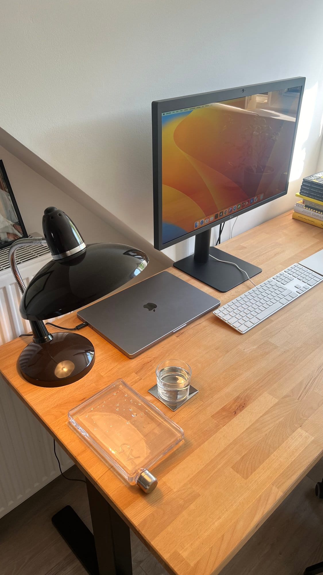 A well-organised home office desk setup featuring an LG UltraFine 5K monitor displaying a macOS desktop, MacBook Pro M1 16″ 2021, Apple Magic Keyboard, black desk lamp, A5 water bottle by memobottle, glass of water on a coaster, and a stack of books