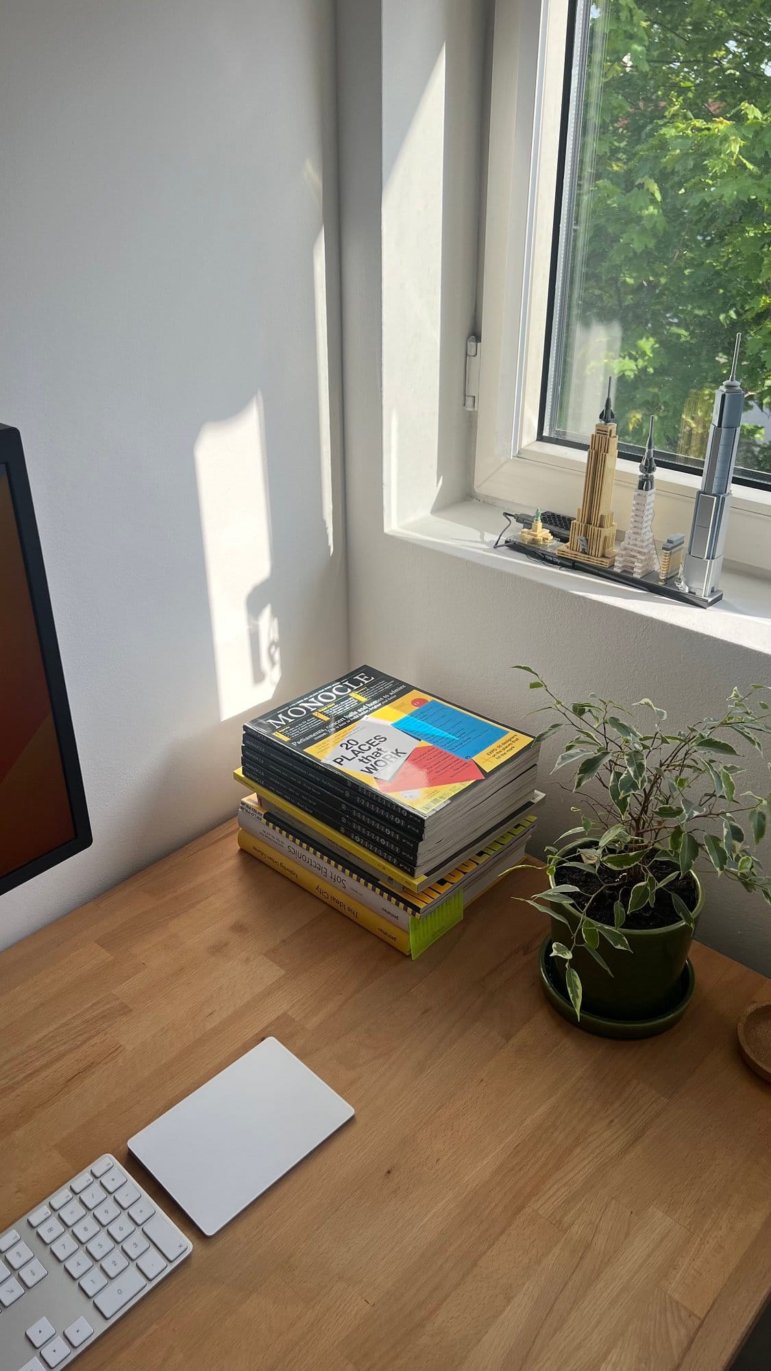 A close-up of a home office desk featuring a stack of books and magazines, a small potted plant, and a LEGO model of famous buildings on the windowsill, all arranged on an IKEA GERTON tabletop
