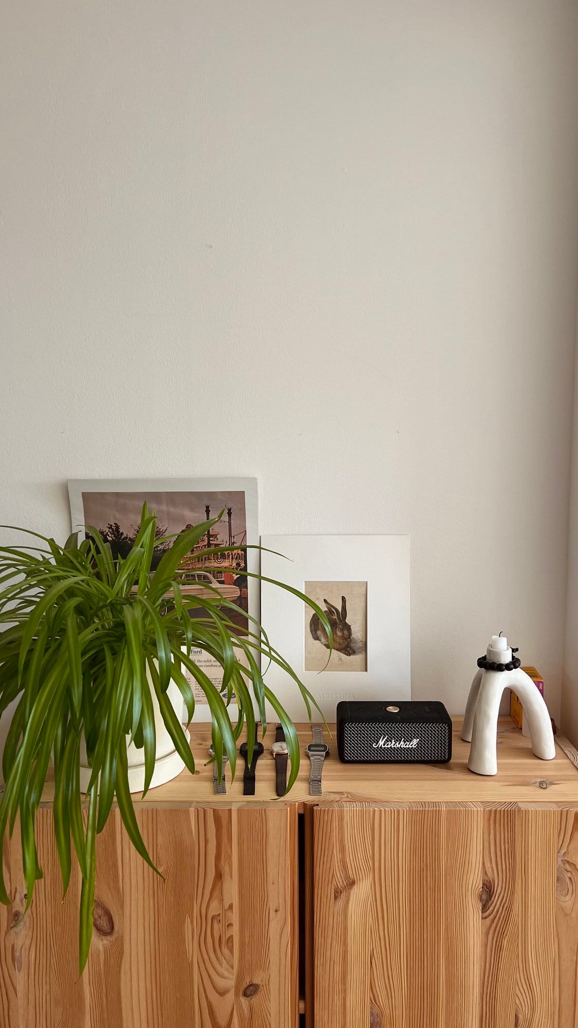 A wooden cabinet topped with a potted spider plant, several smartwatch bands, a Marshall portable speaker, a candle holder, and framed art prints leaning against the wall