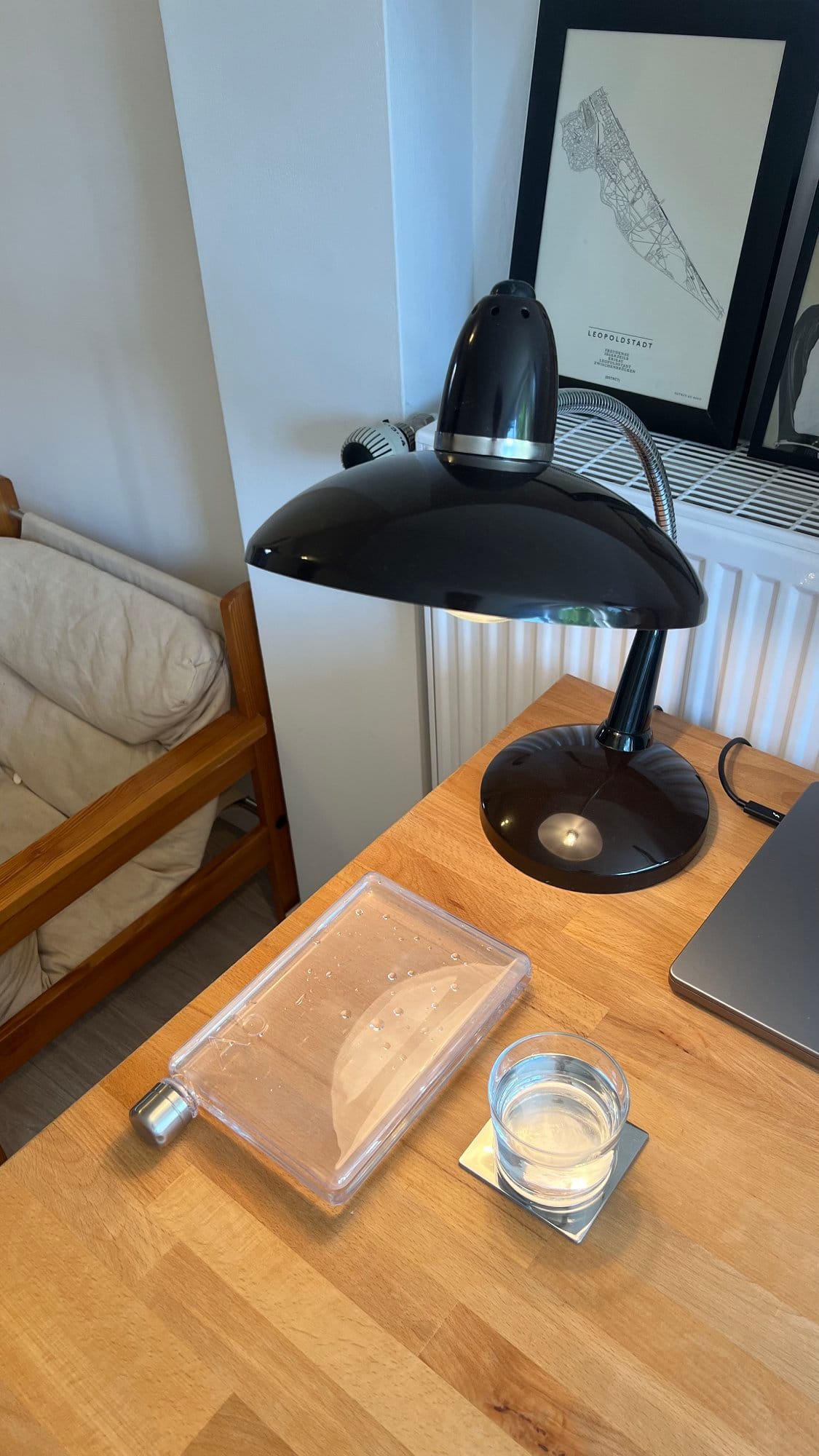 A close-up of a home office desk featuring a vintage black desk lamp, a clear A5 memobottle water bottle, a glass of water on a coaster, part of a MacBook Pro M1 16″ 2021, and a framed map of Vienna’s Leopoldstadt district in the background
