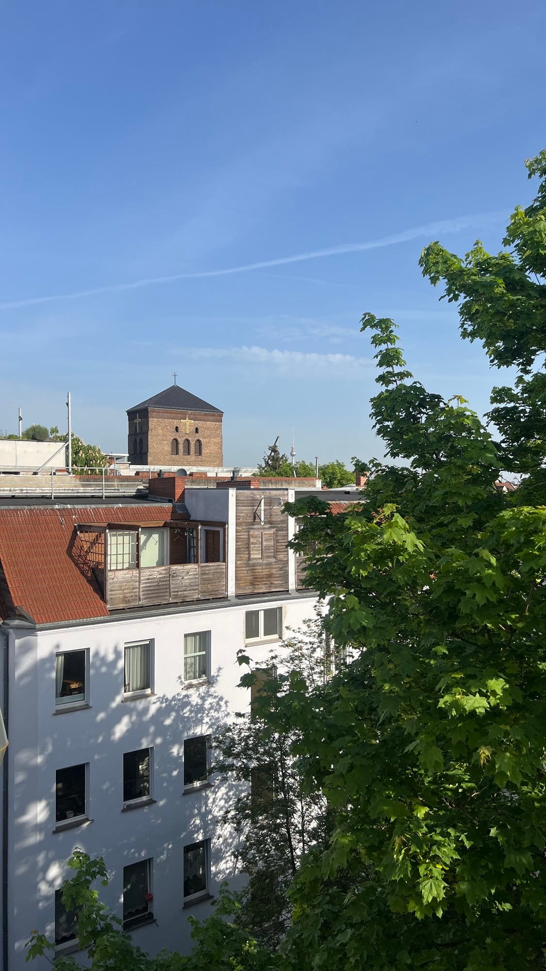 A view of a residential area in Berlin with a white building featuring multiple windows and balconies, a large leafy tree, and a historic brick tower with crosses and a pitched roof, all set against a clear blue sky