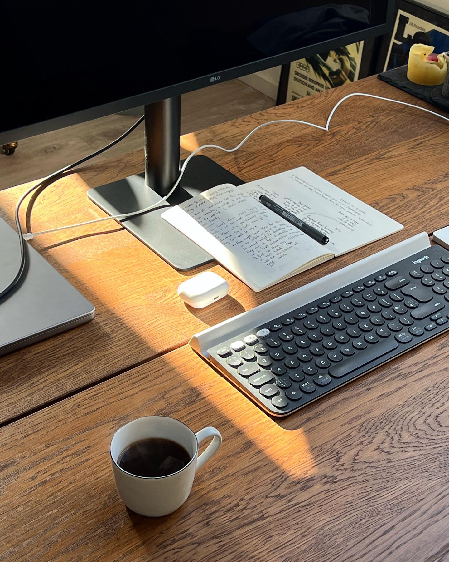 A close-up of a home office desk featuring a Logitech K780 keyboard, AirPods Pro 2021, a notebook with handwritten notes, a pen, and a cup of black coffee