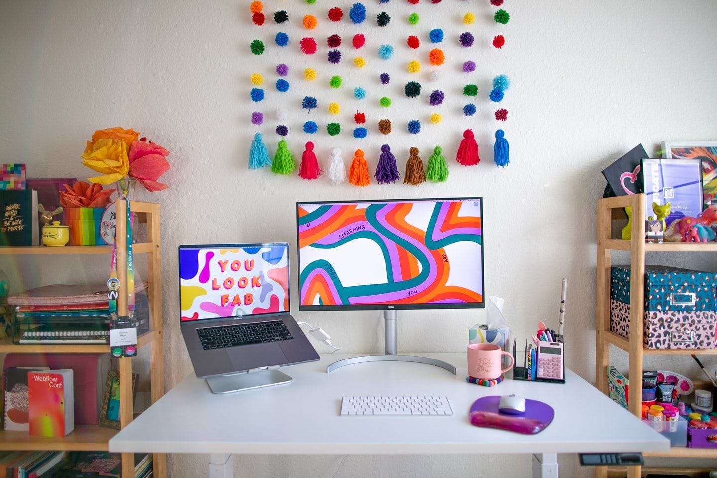 A colourful desk setup with a laptop, monitor, keyboard, mouse, and various decorative items, including paper flowers, a rainbow tassel wall hanging, and shelves filled with books and office supplies