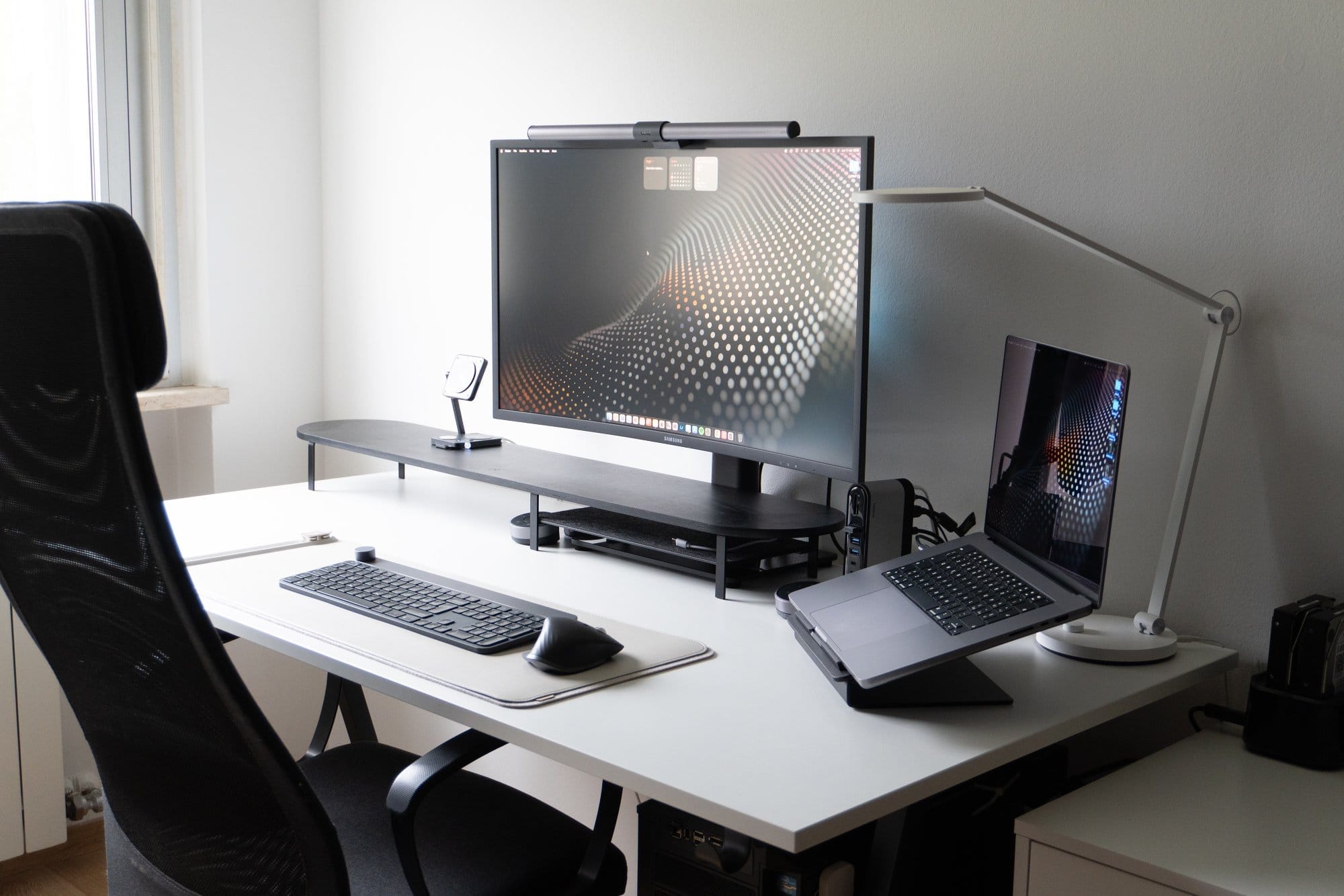 A modern desk setup with a large monitor, laptop on a stand, keyboard, mouse, desk lamp, and ergonomic chair