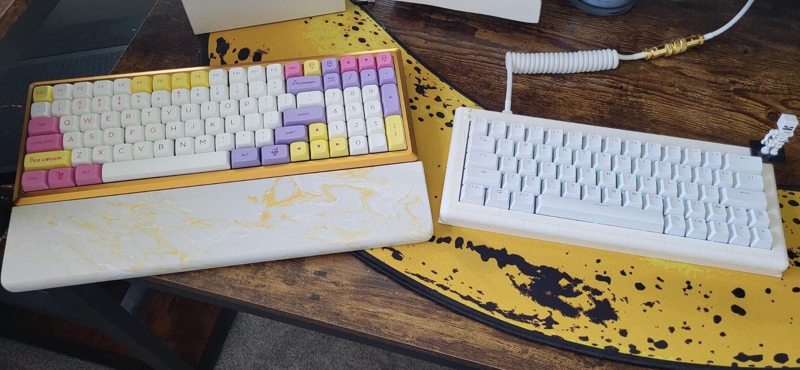 A desk setup featuring two mechanical keyboards; the left keyboard has a gold case with pastel NP PBT Ice Cream keycaps, and the right keyboard is white with YMDK Double Shot Shine Through keycaps