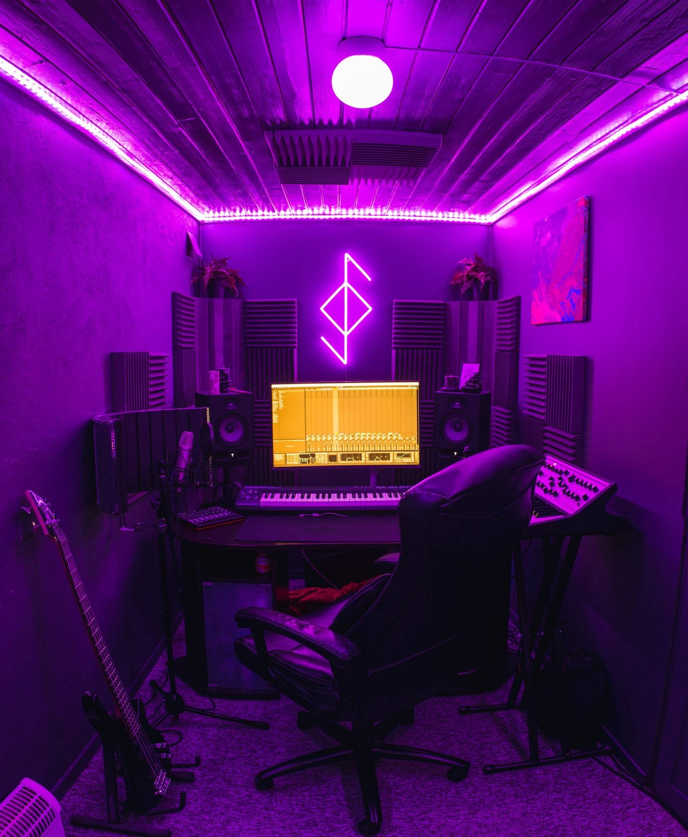A vibrant and cosy home music studio with purple LED lighting, featuring a sleek black desk equipped with a computer monitor, keyboard, speakers, and recording equipment