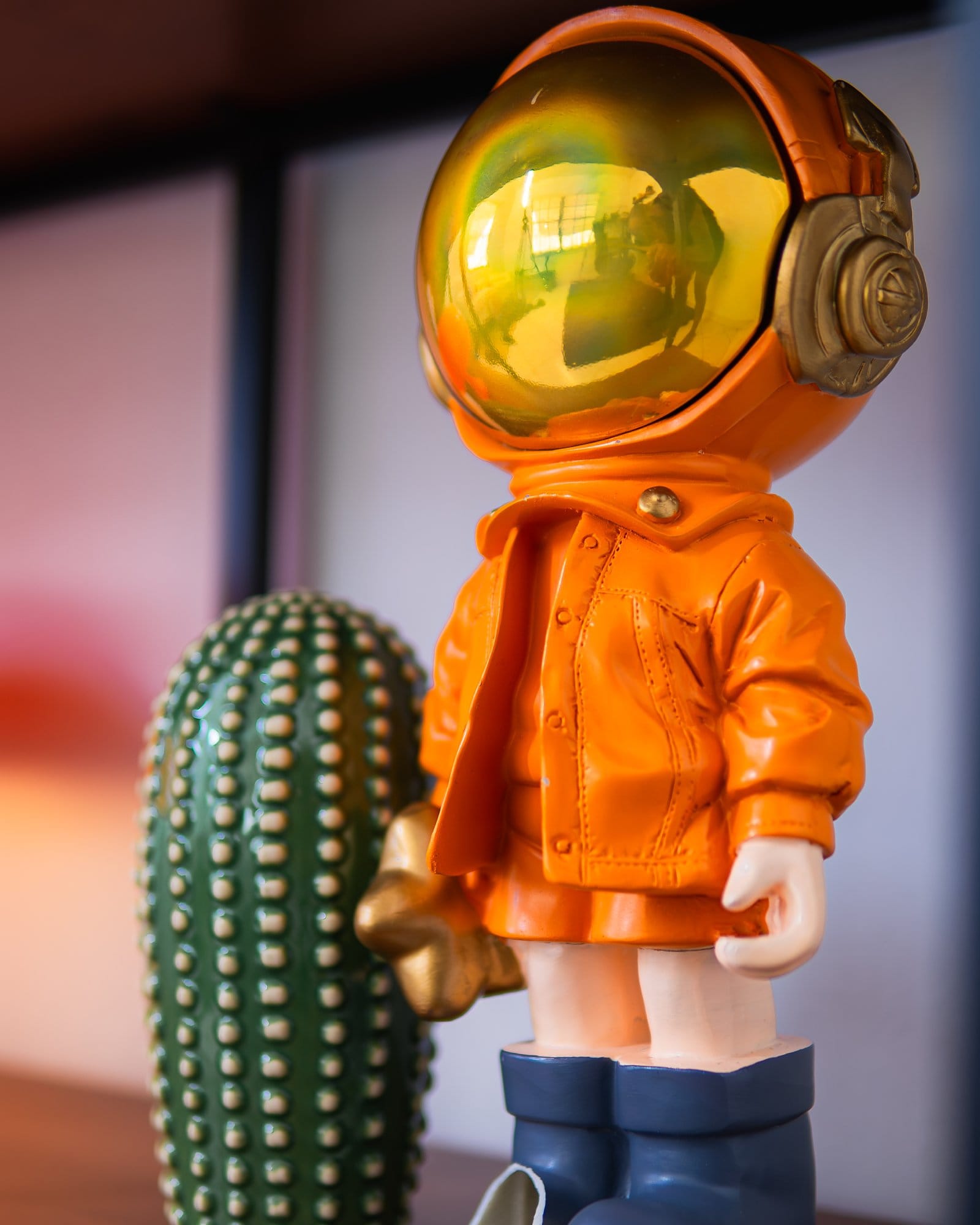 A close-up of a stylised astronaut figurine in an orange suit with a reflective gold visor, standing next to a green cactus decor piece