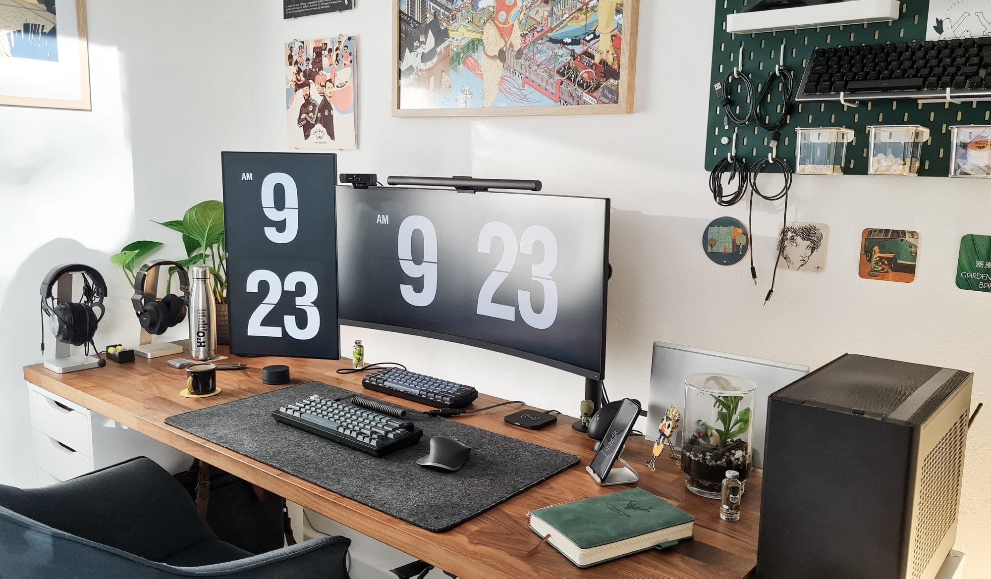A well-organised desk setup featuring dual monitors displaying a large clock, two mechanical keyboards, and a Logitech mouse on a grey felt desk mat