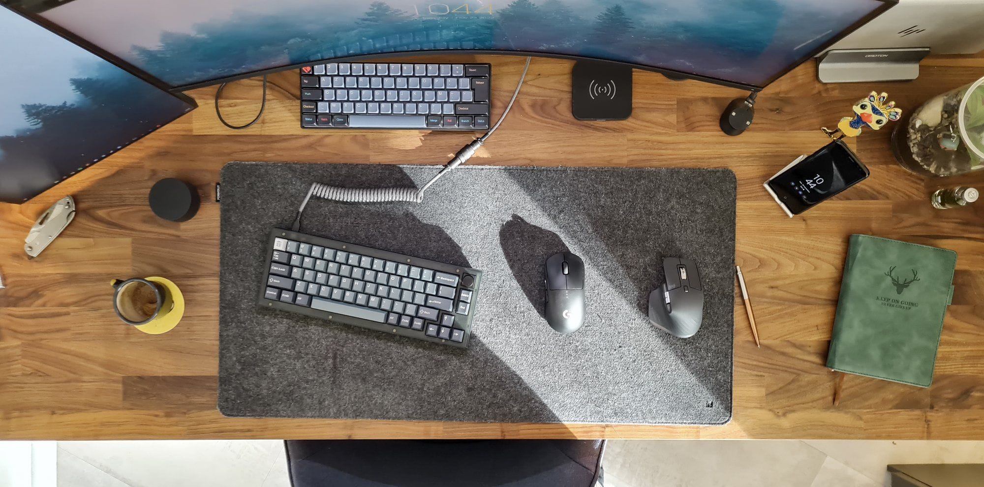 A top-down view of a desk setup featuring two mechanical keyboards, two Logitech mice on a grey felt desk mat, with a cup of coffee, smartphone, and green notebook arranged neatly on a wooden desk