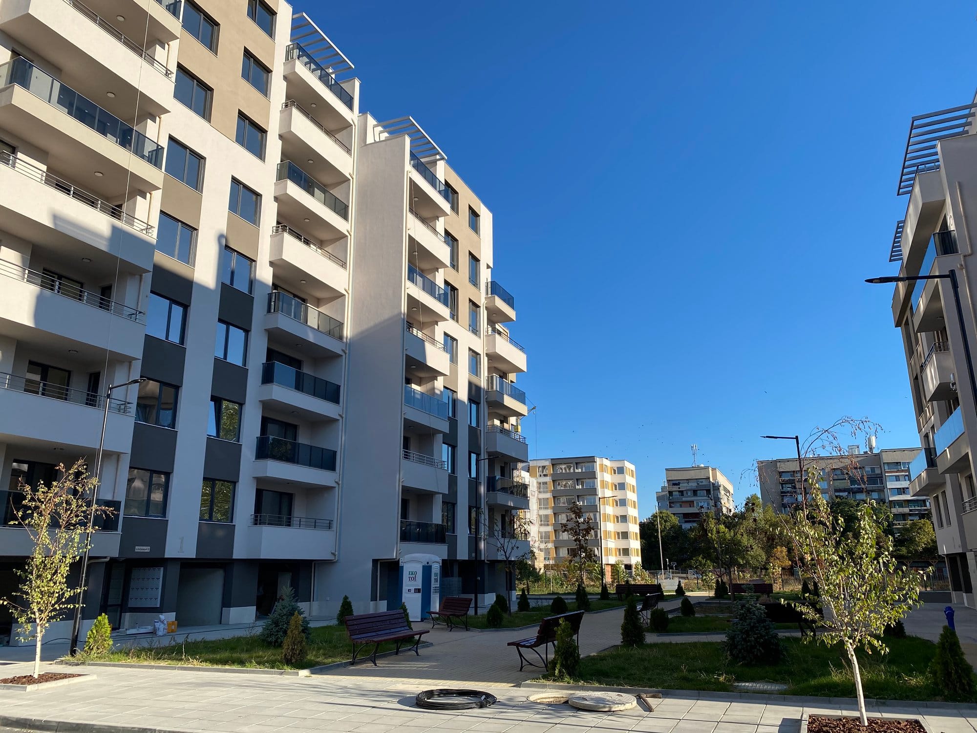 A sunny day in a modern residential complex with multi-storey apartment buildings, landscaped green areas, benches, and young trees in Plovdiv, Bulgaria