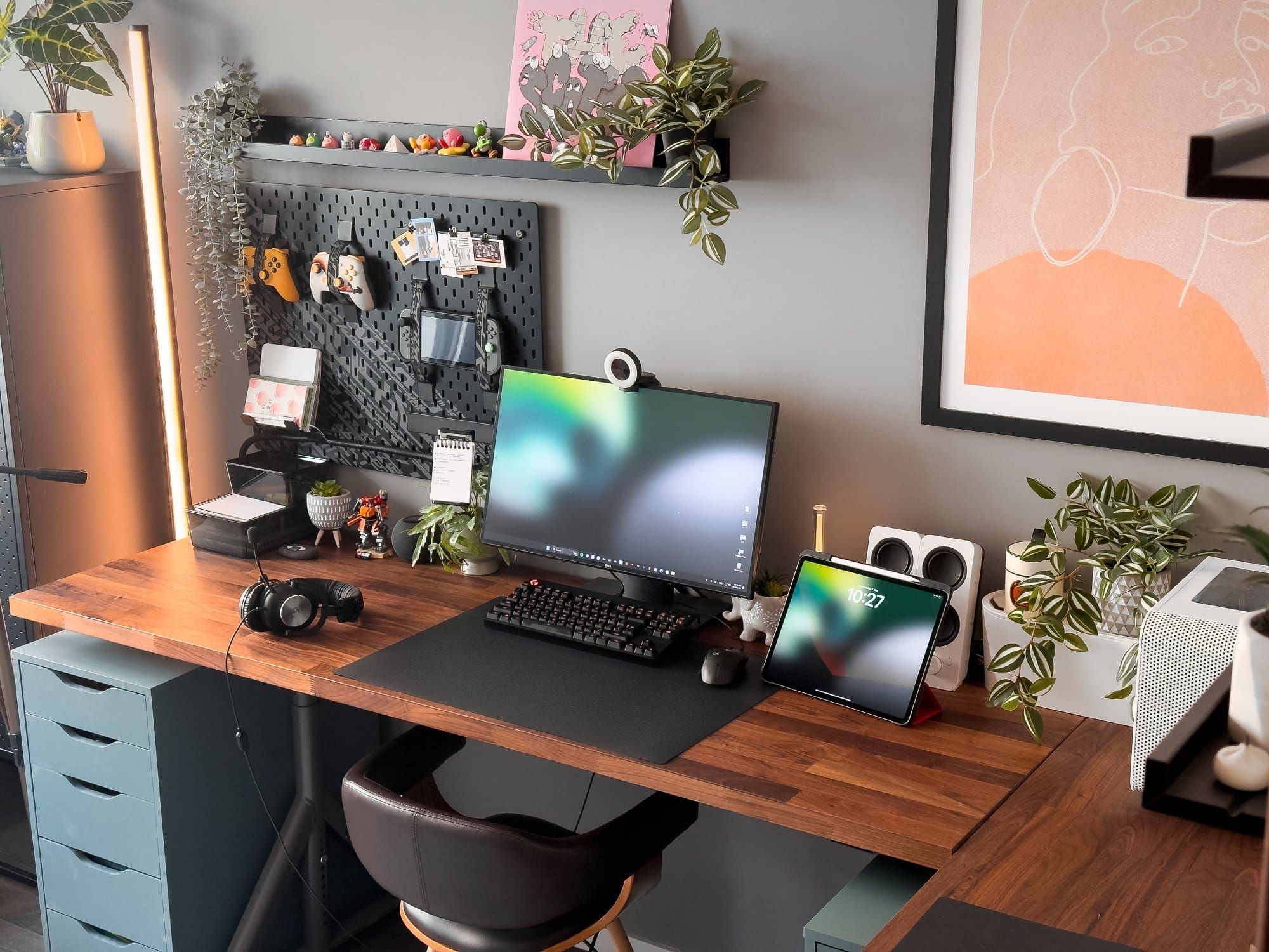 A modern and neatly arranged workspace featuring a Dell monitor with a Razer Kiyo webcam, Logitech G Pro keyboard and mouse, an iPad Pro 12.9 (3rd Gen), and Logitech G Pro X headphones on a wooden IKEA KARLBY desk with IKEA ALEX drawers, complemented by plants, framed artwork, and an IKEA UPPSPEL pegboard