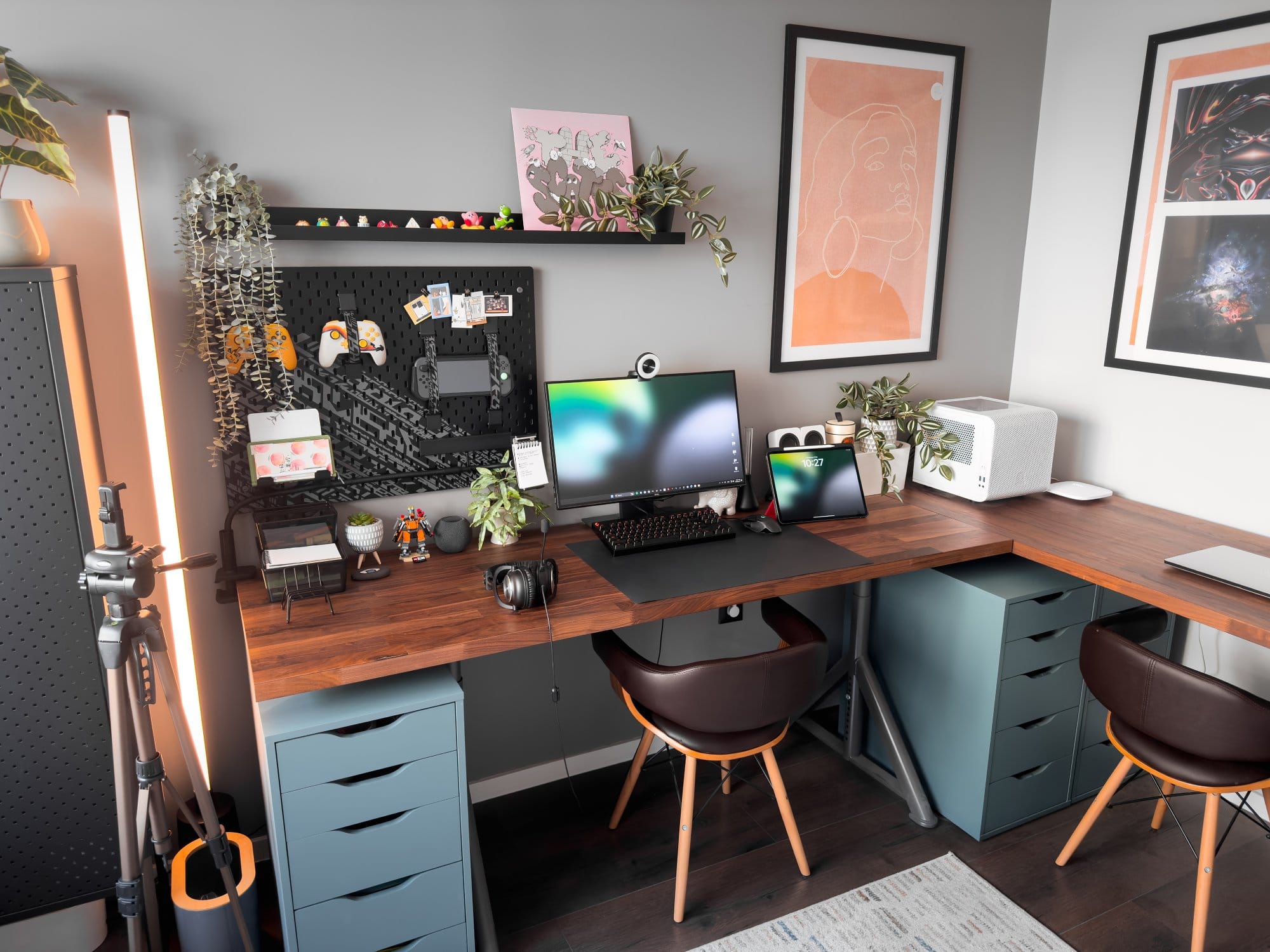 An L-shaped home office setup featuring a Dell monitor with Razer Kiyo webcam, Logitech G Pro keyboard and mouse, an iPad Pro 12.9 (3rd Gen), Logitech G Pro X headphones, and a printer, all arranged on a wooden IKEA KARLBY desk with IKEA ALEX drawers, complemented by plants, artwork, and an IKEA UPPSPEL pegboard
