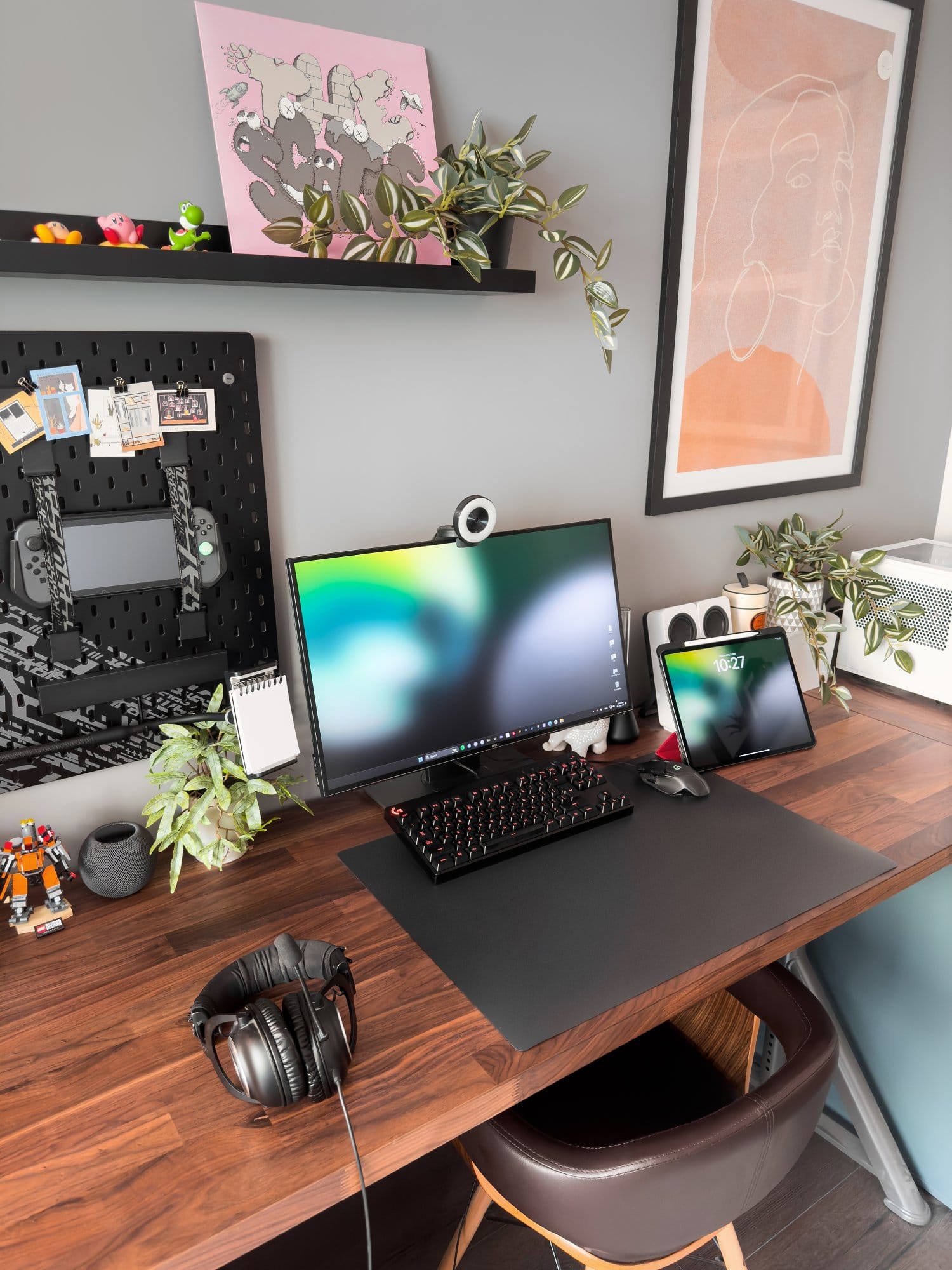 A home office setup with a Dell monitor and Razer Kiyo webcam, Logitech G Pro keyboard and mouse, an iPad Pro 12.9 (3rd Gen), and Logitech G Pro X headphones on a wooden IKEA KARLBY desk, decorated with plants, framed artwork, and an IKEA UPPSPEL pegboard in the background