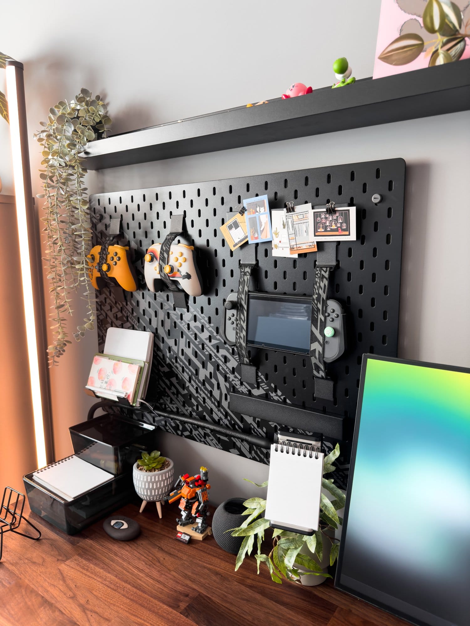 A close-up view of a workspace featuring an IKEA UPPSPEL pegboard with controllers, notes, and a Nintendo Switch, a variety of plants and decorations, and a Dell monitor partially visible, all set on a wooden IKEA KARLBY desk