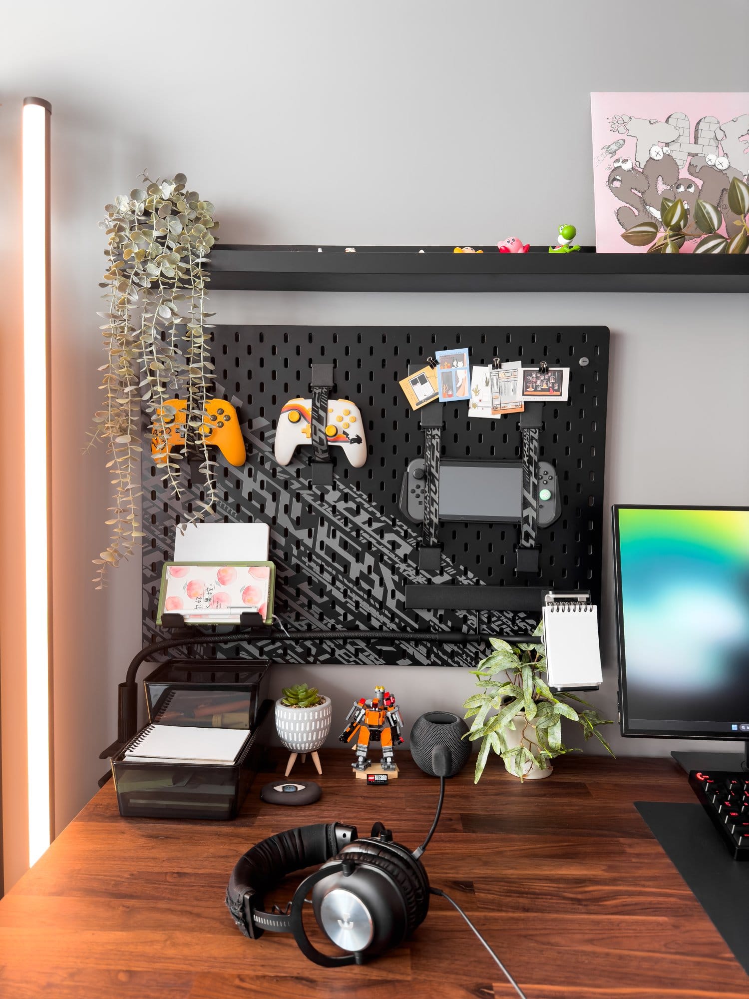 A close-up of a modern desk setup featuring a Dell monitor, Logitech G Pro X headphones, and an IKEA UPPSPEL pegboard holding controllers, notes, and a Nintendo Switch, alongside plants and decorative items, all arranged on a wooden IKEA KARLBY desk