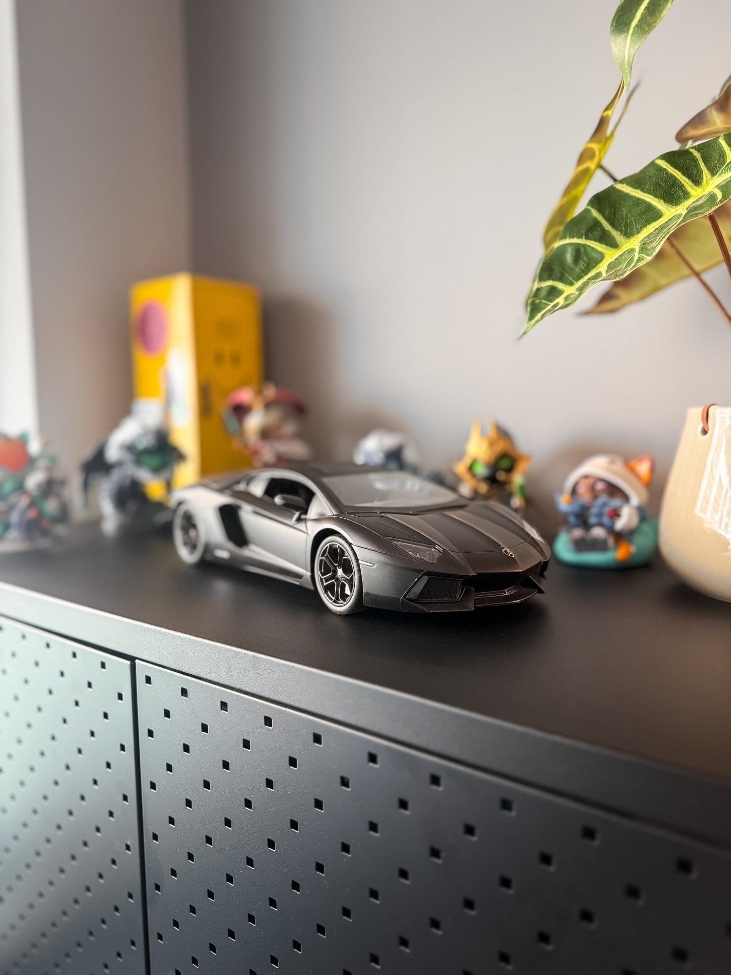 A close-up of a black perforated shelf displaying a model car, surrounded by various small figurines and a plant in a ceramic pot