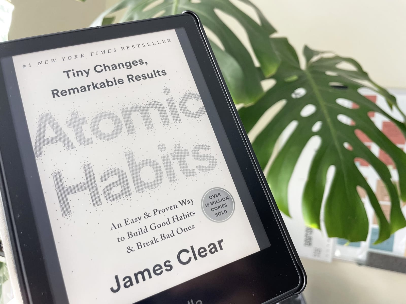 A close-up of an e-reader displaying the book “Atomic Habits” by James Clear, set against the backdrop of a leafy plant