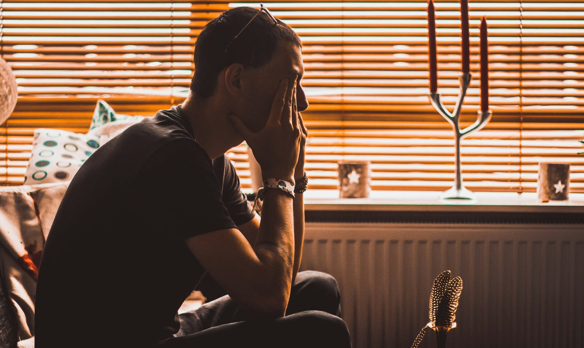 A tired man sitting with his hands on his face in a dimly lit room, with window blinds casting a warm, golden light