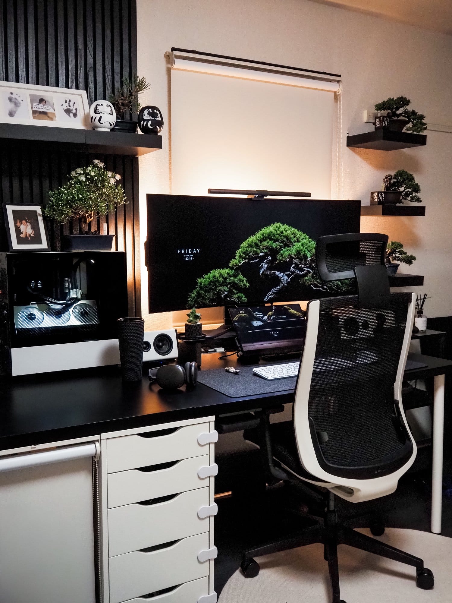 A modern desk setup featuring a Corsair monitor with a BenQ light bar, a MacBook Pro laptop, and a white AIMchair gaming chair, decorated with bonsai trees and Daruma dolls